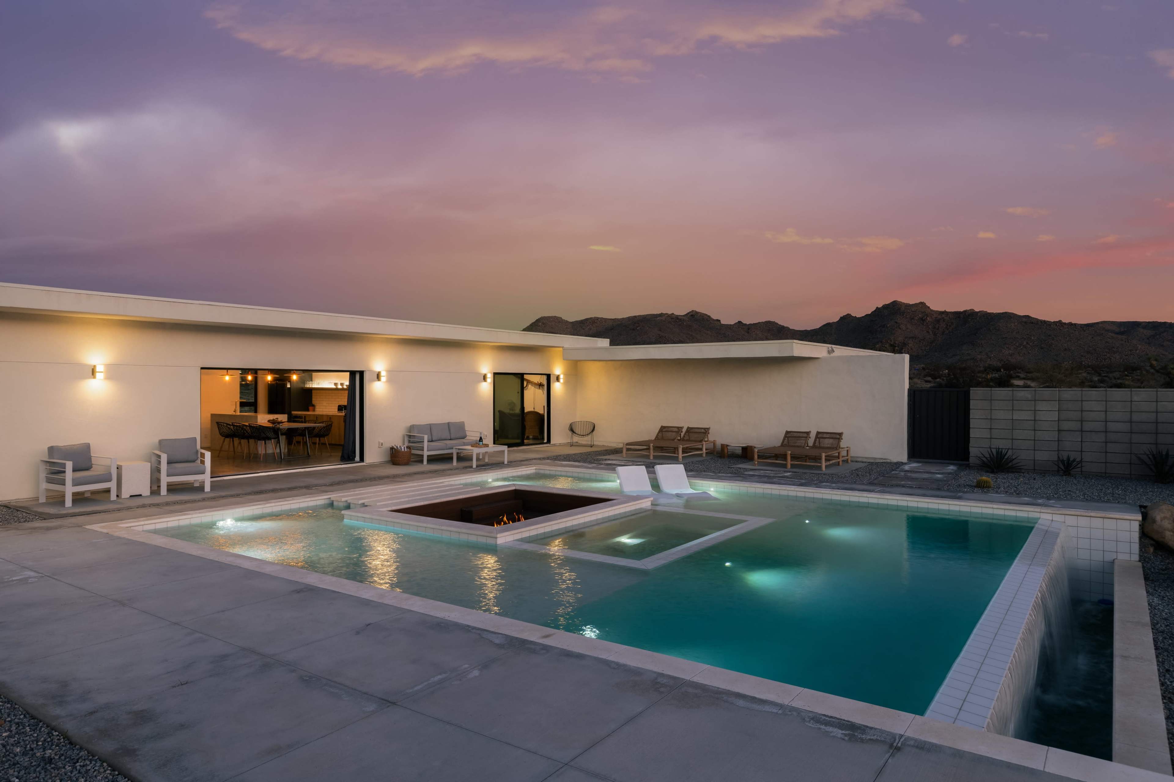 A modern outdoor pool is illuminated at dusk, featuring a sunken hot tub and surrounded by minimalistic seating and desert mountains in the background.