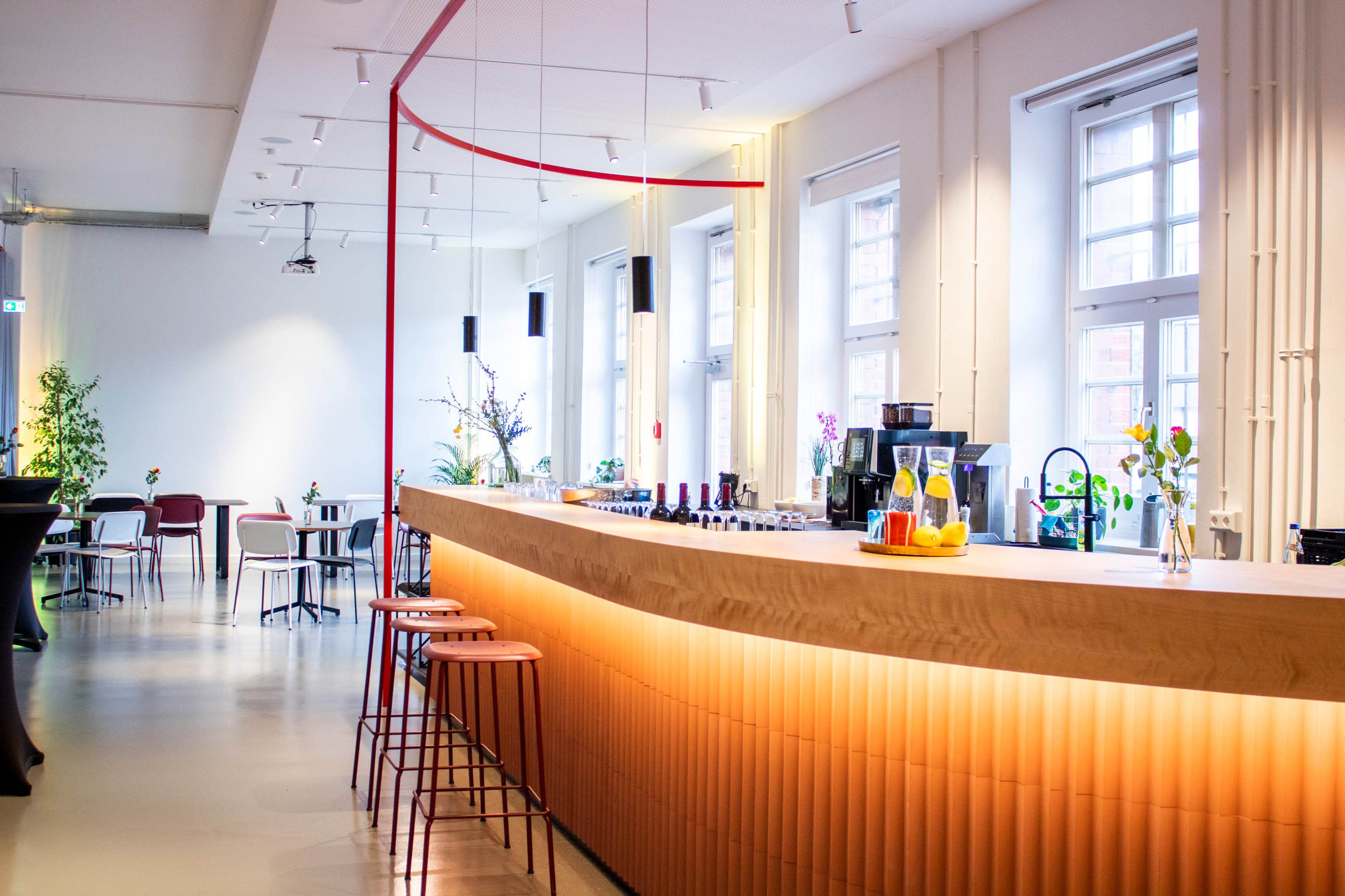 A modern, spacious café with a light wooden bar, colorful stools, and large windows allowing natural light to fill the room.