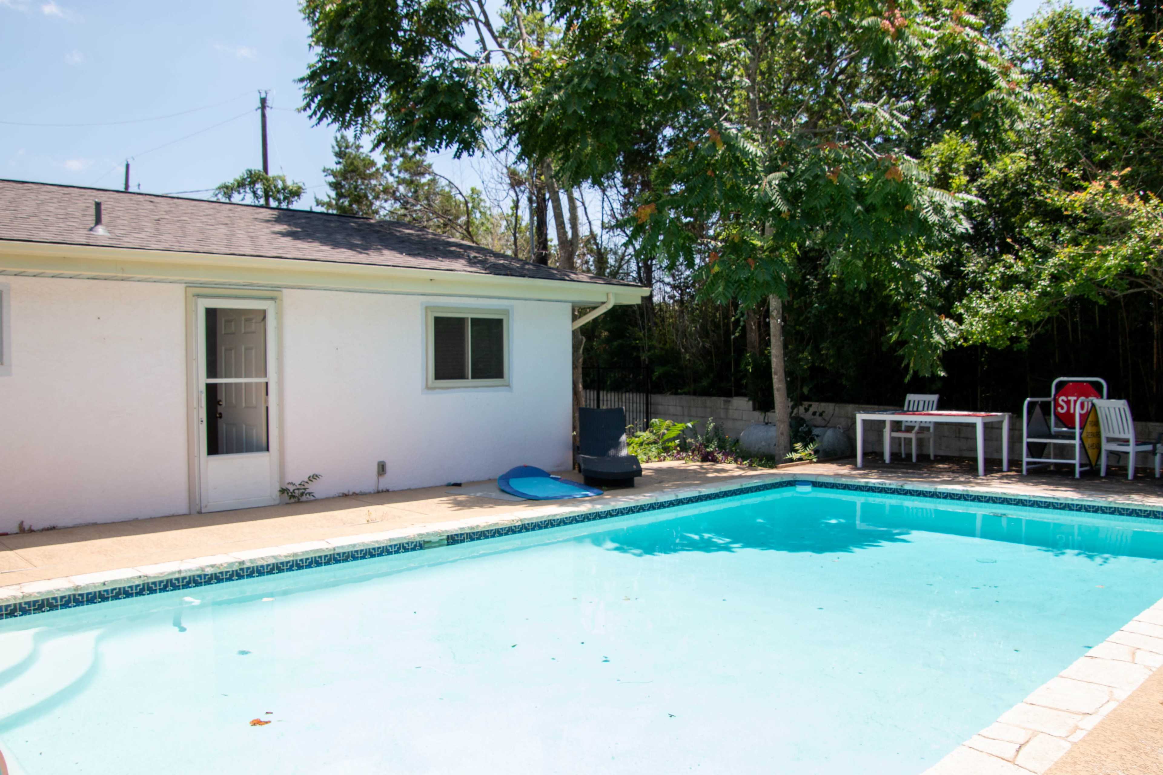 A clear swimming pool is located beside a white house with a door leading to an outdoor area, surrounded by trees and patio furniture.