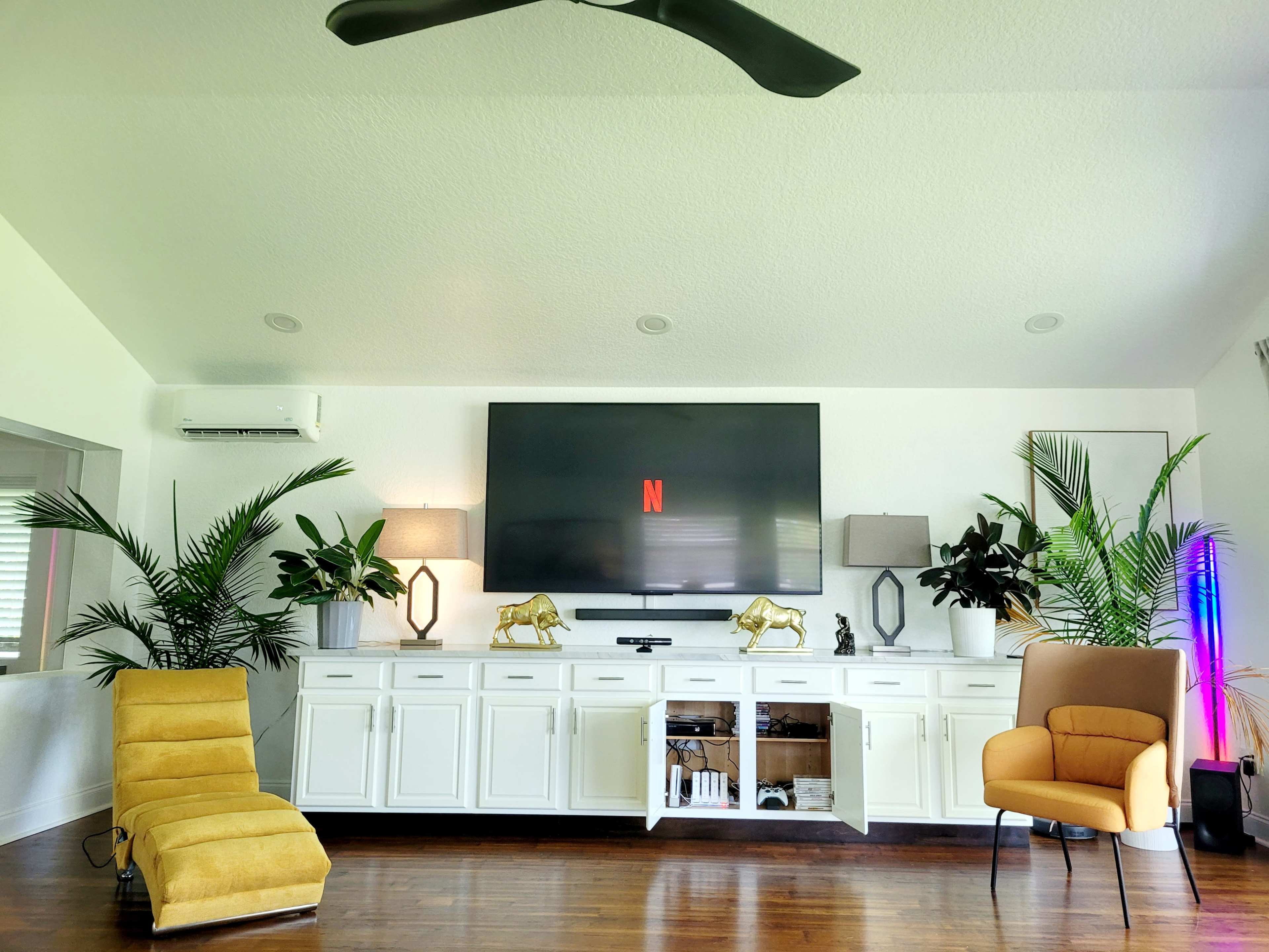 A modern living room features a large TV mounted on the wall above a white cabinet, flanked by plants and lamps, with yellow chairs on either side.