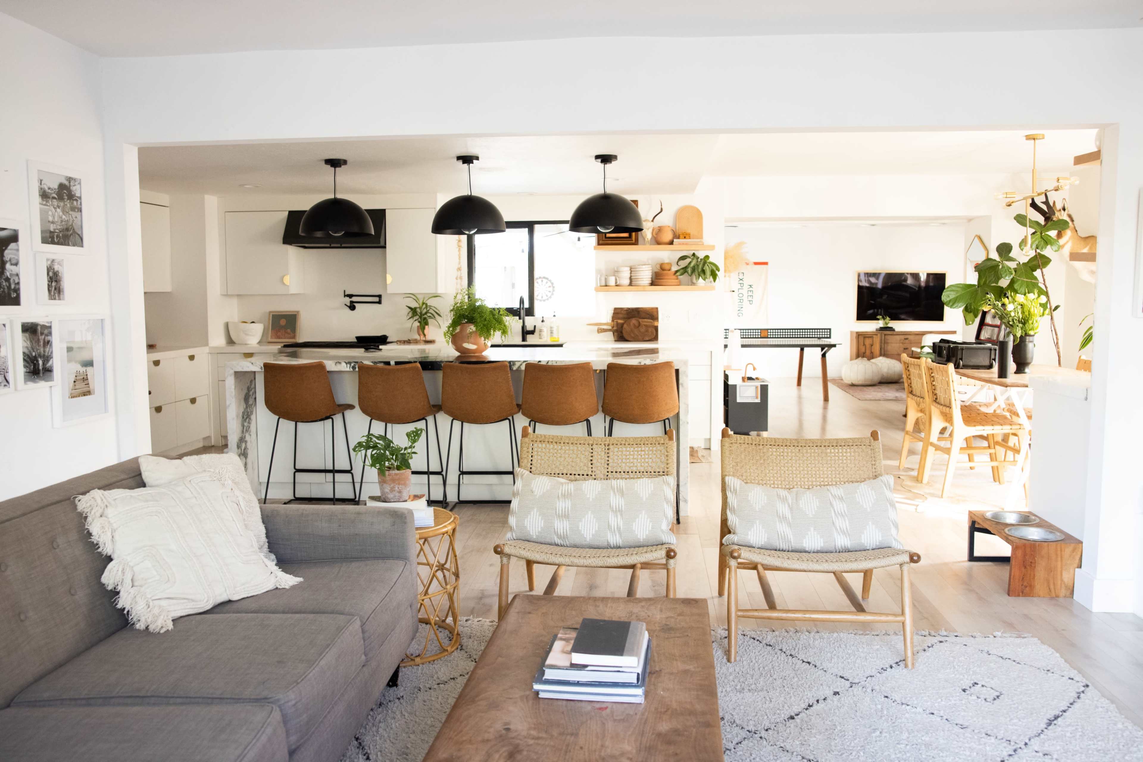 A modern open-concept living space features a gray sofa, a wooden coffee table, and rattan chairs facing a kitchen with a large island and bar seating.