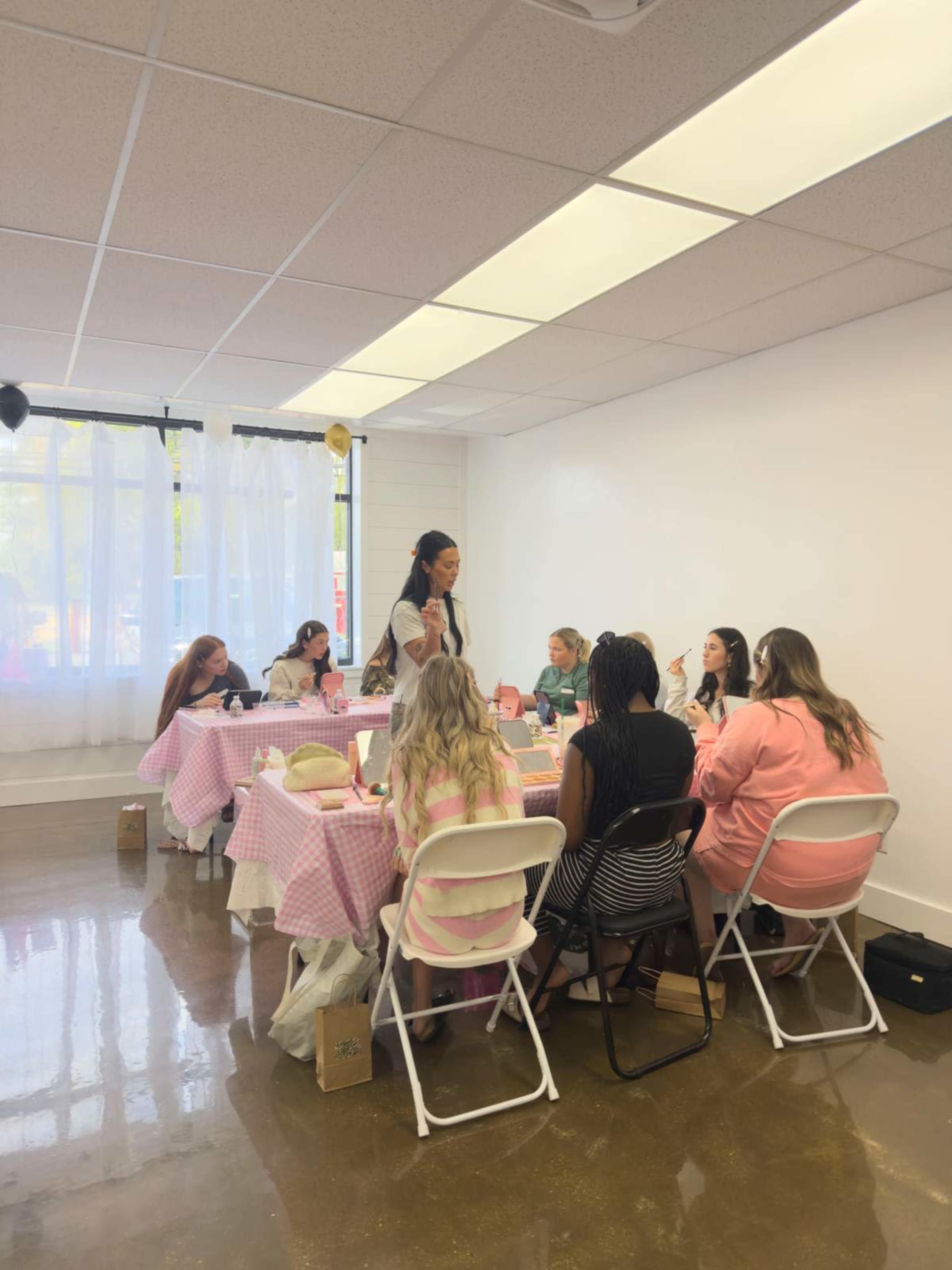 A group of women is seated around a table covered with a pink checkered cloth in a brightly lit room, engaged in a craft activity.