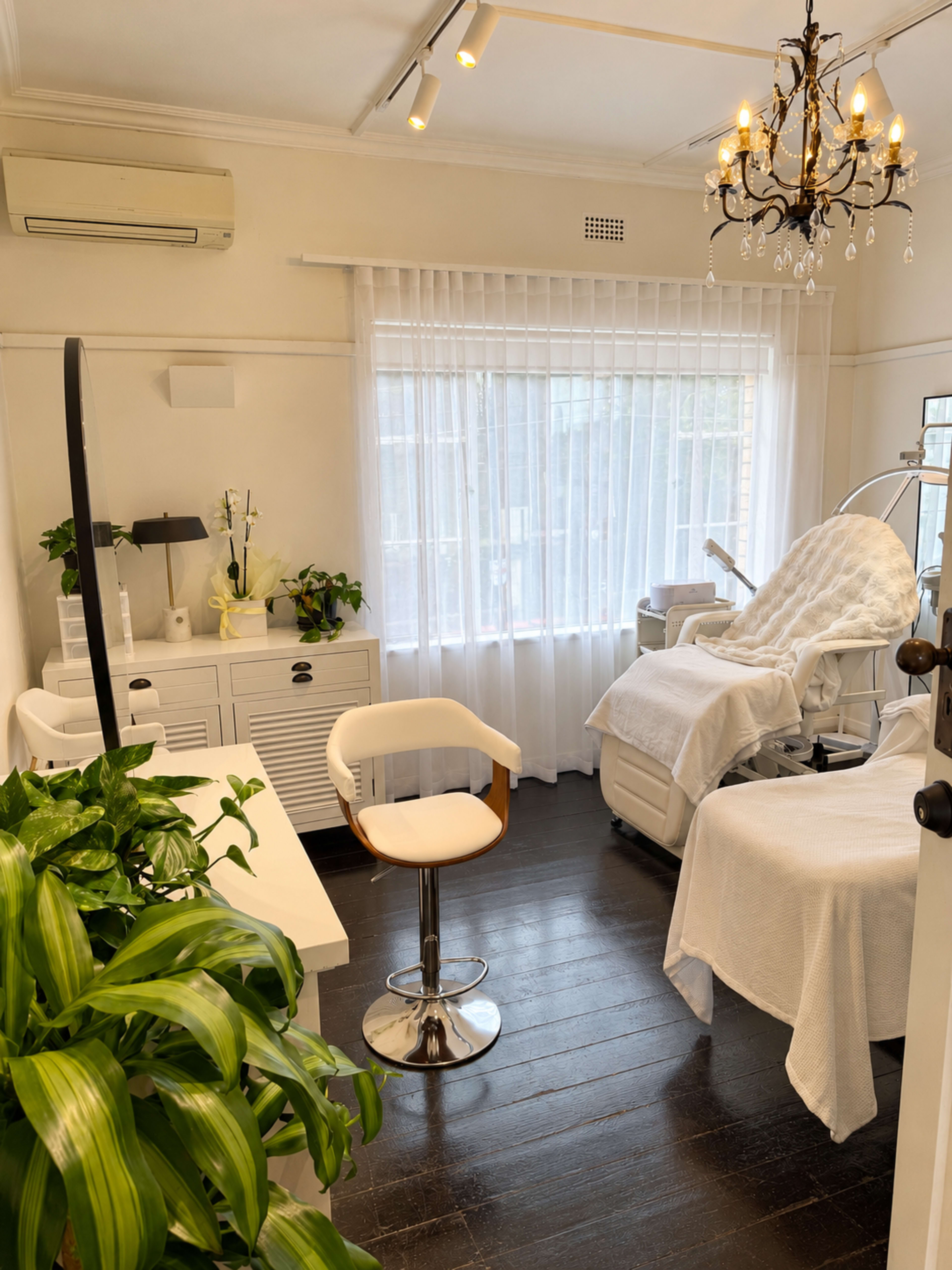 The image shows a well-lit treatment room with a treatment bed, a white chair, a desk, and a window adorned with sheer curtains.