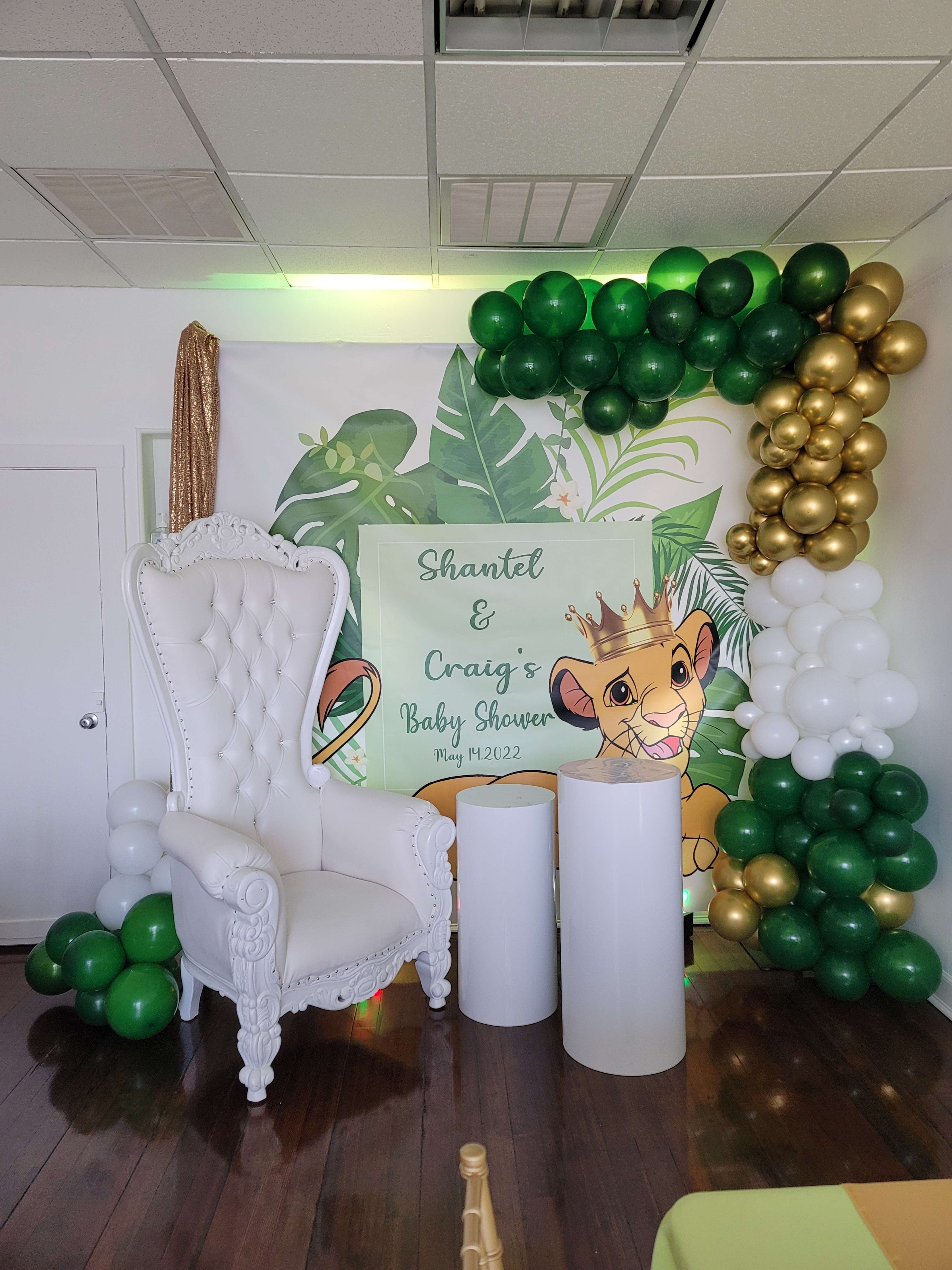 The image shows a decorative backdrop for a baby shower featuring a white throne chair, green and gold balloons, and a sign with the names "Shantel & Craig" along with a Lion King theme.