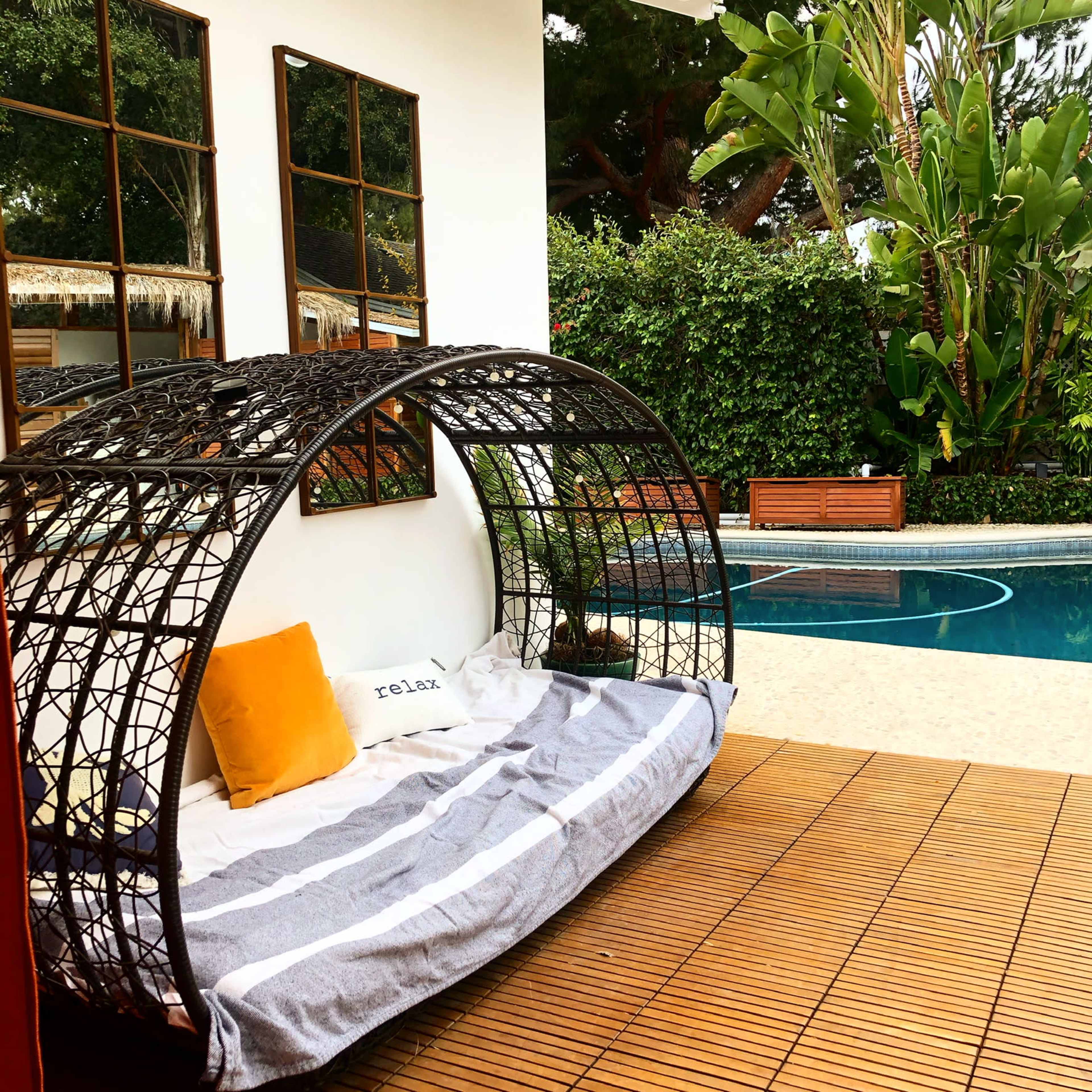 A curved outdoor seating area made of woven material beside a swimming pool, surrounded by greenery and mirrors on the wall.