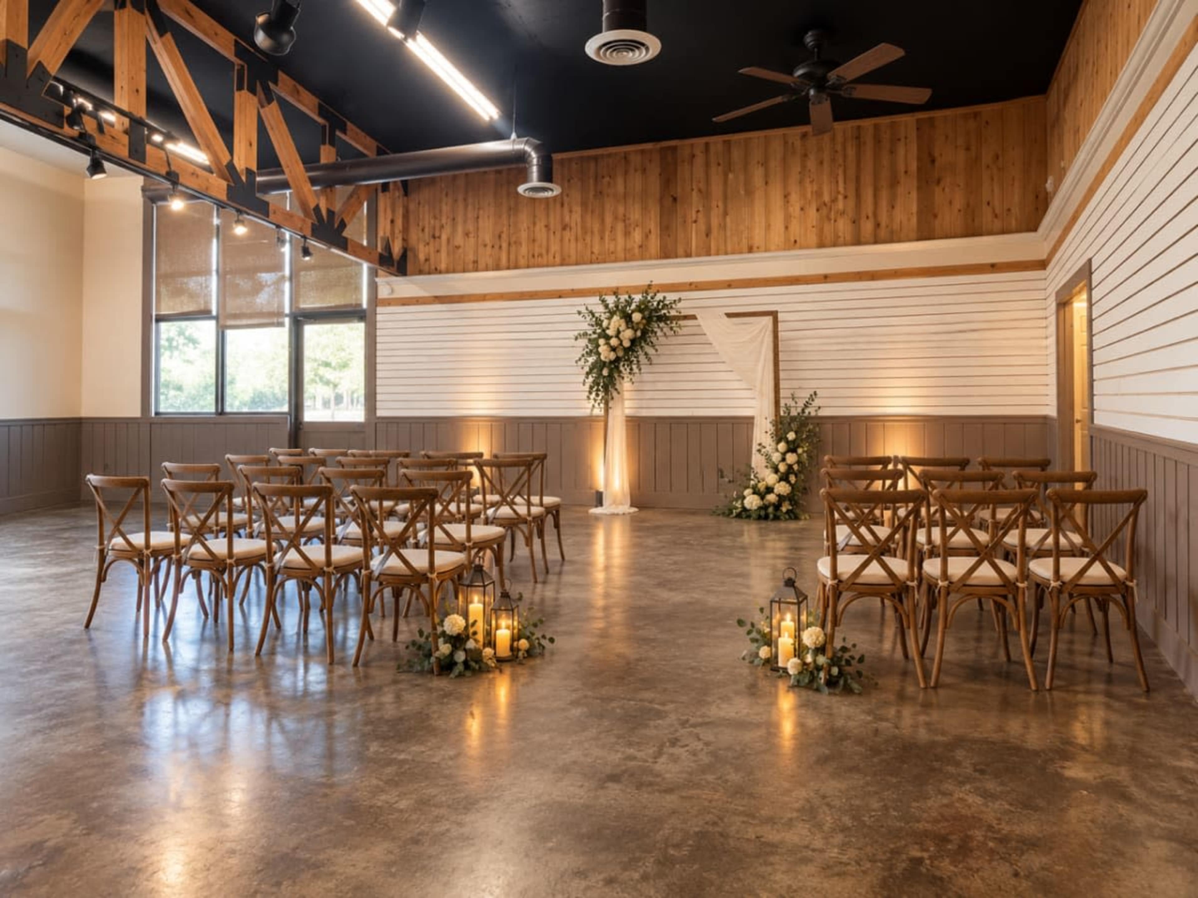 A wedding ceremony setup features rows of wooden chairs facing a floral arch, with decorative lanterns and plants on the floor in a spacious, well-lit venue.