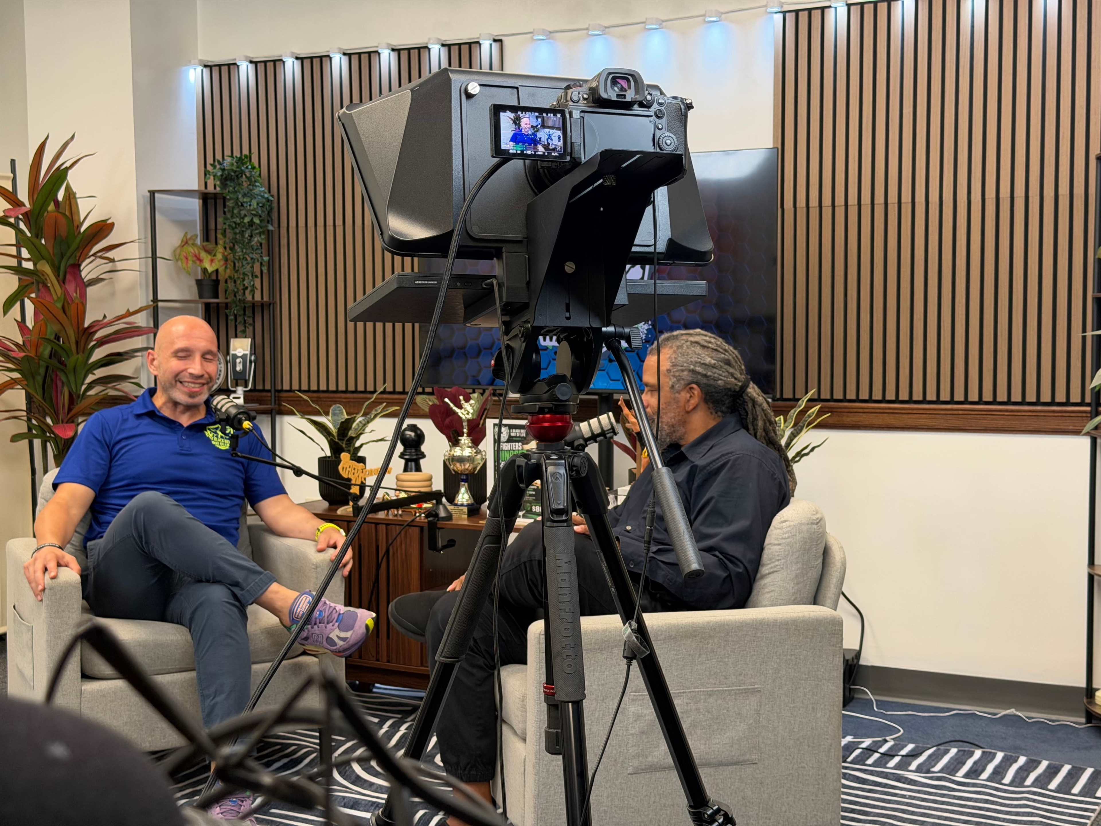 Two men are seated in a studio, with one man on the left smiling and the other speaking, while a camera is positioned to capture the conversation.