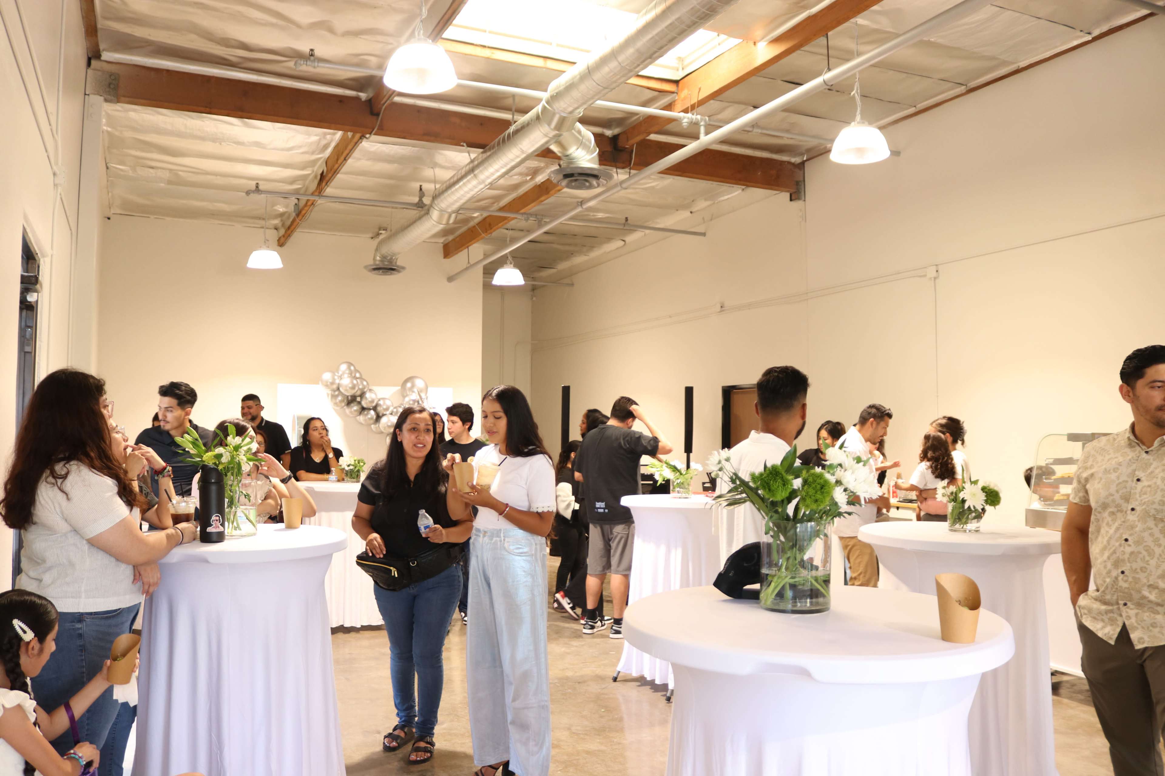 A spacious indoor event space features guests mingling around several tall tables decorated with flowers and refreshments.