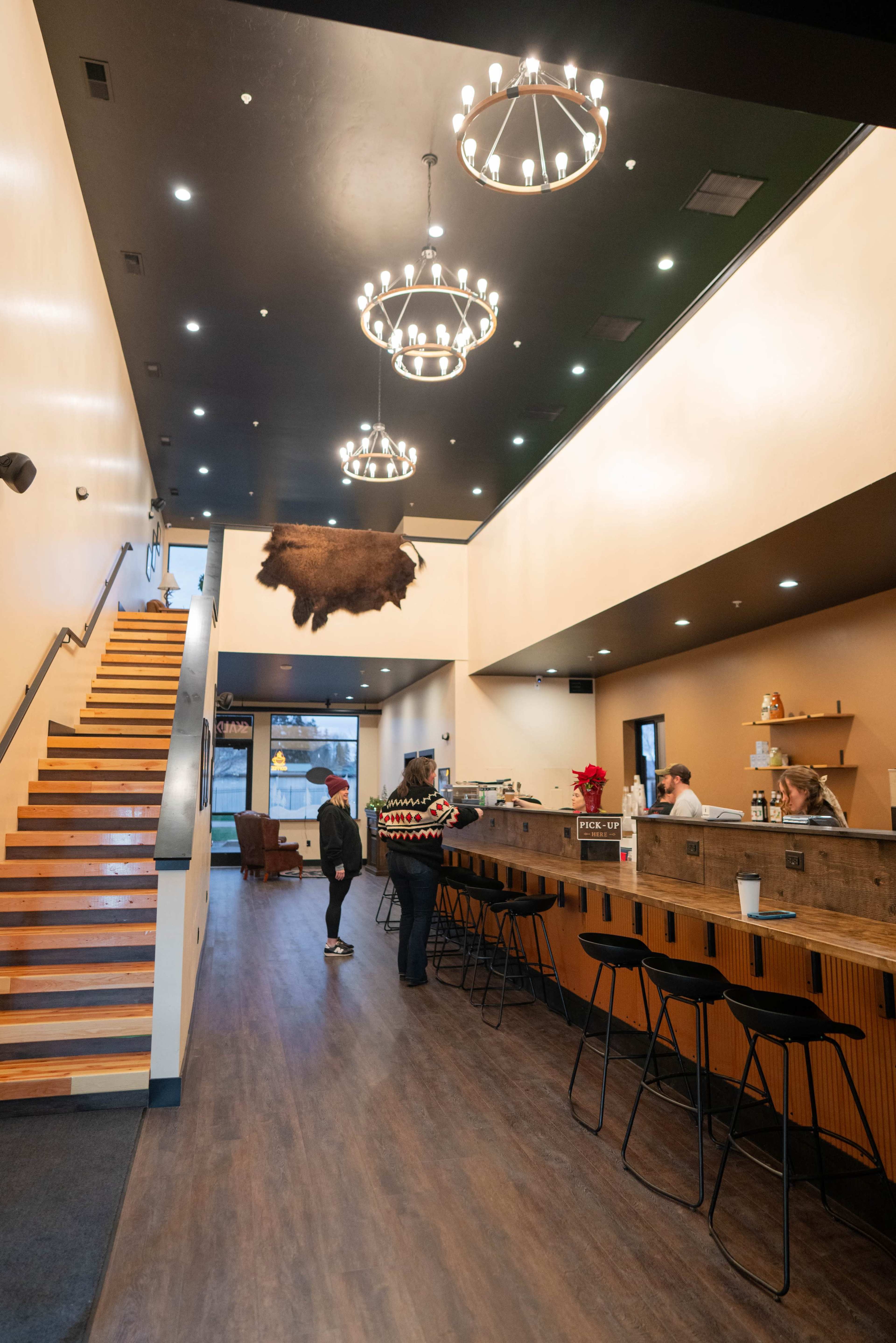 The image shows a modern café with a wooden counter, a staircase on one side, and a mounted decorative animal hide hanging from the ceiling.
