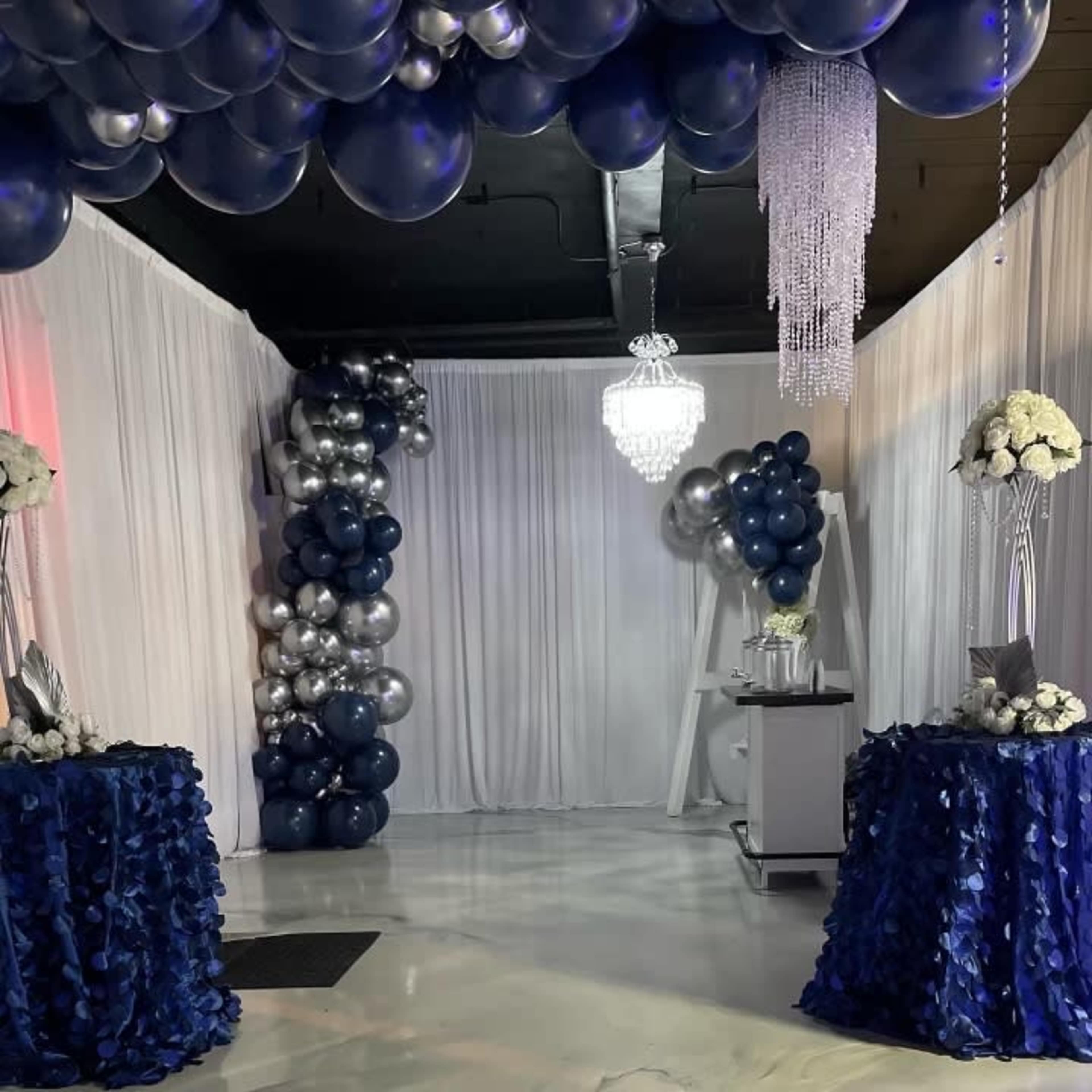 The image shows a decorated event space featuring a balloon arch, elegant tables with floral arrangements, and a chandelier overhead.