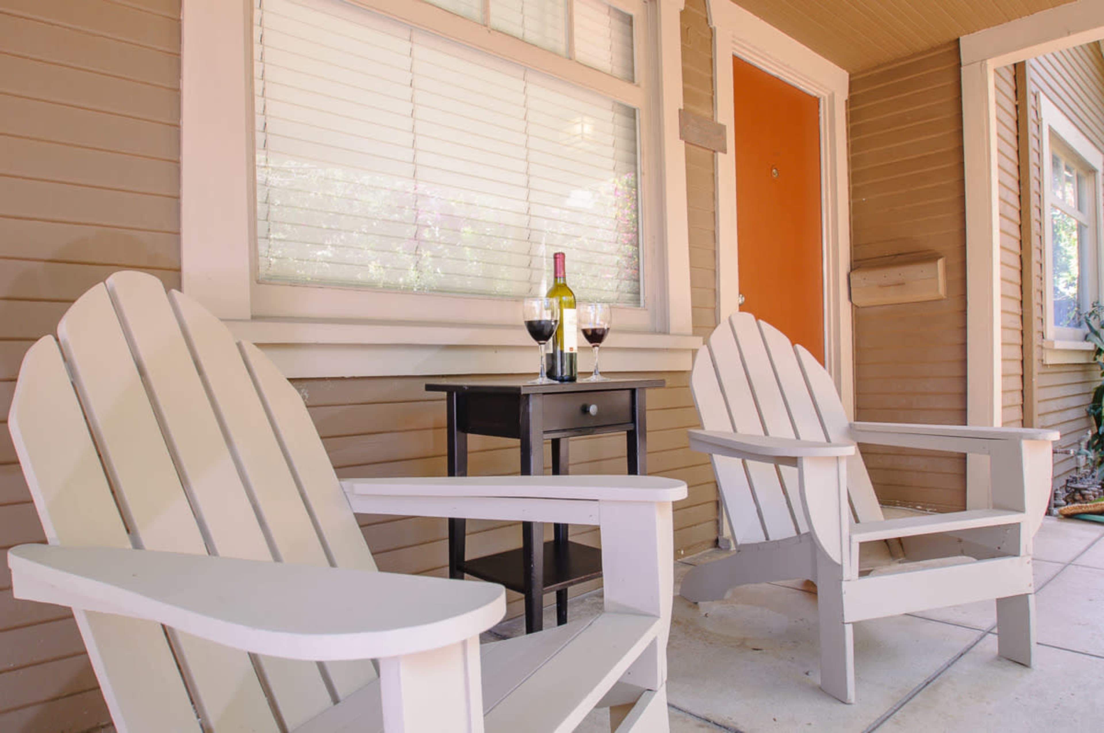 Two adirondack chairs sit on a porch beside a small table holding a bottle of wine and two glasses.
