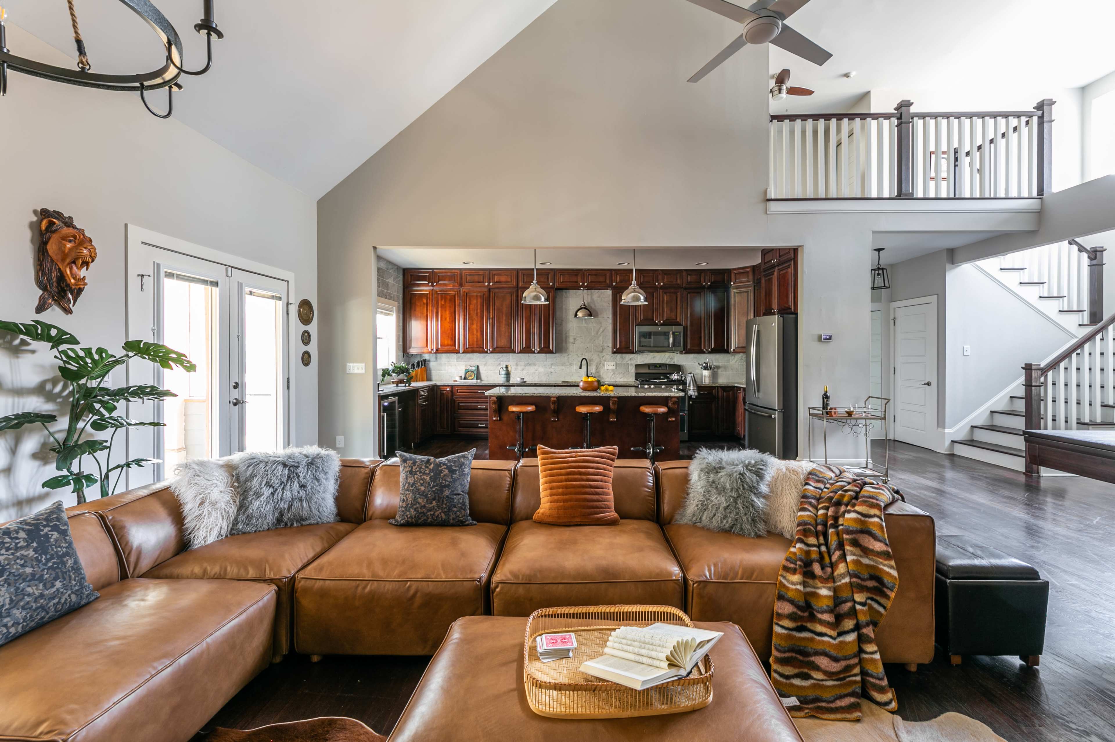 A spacious living room features a large brown leather sectional sofa, an open kitchen with dark wood cabinetry, and a staircase complemented by white railings.
