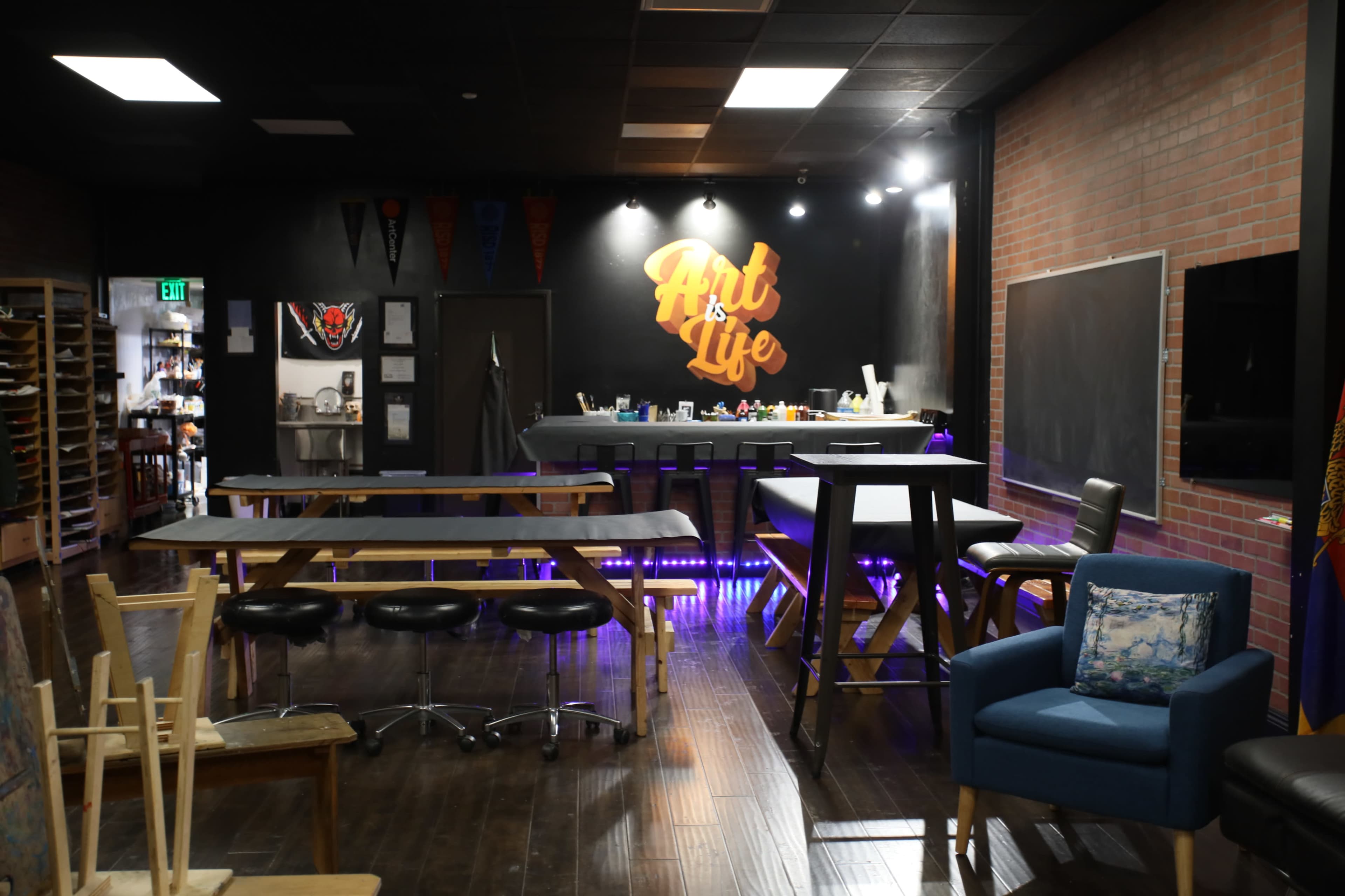 The image shows a modern art studio featuring a bar-like workspace, tables, and various seating options with a colorful "Art is Life" mural on the wall.