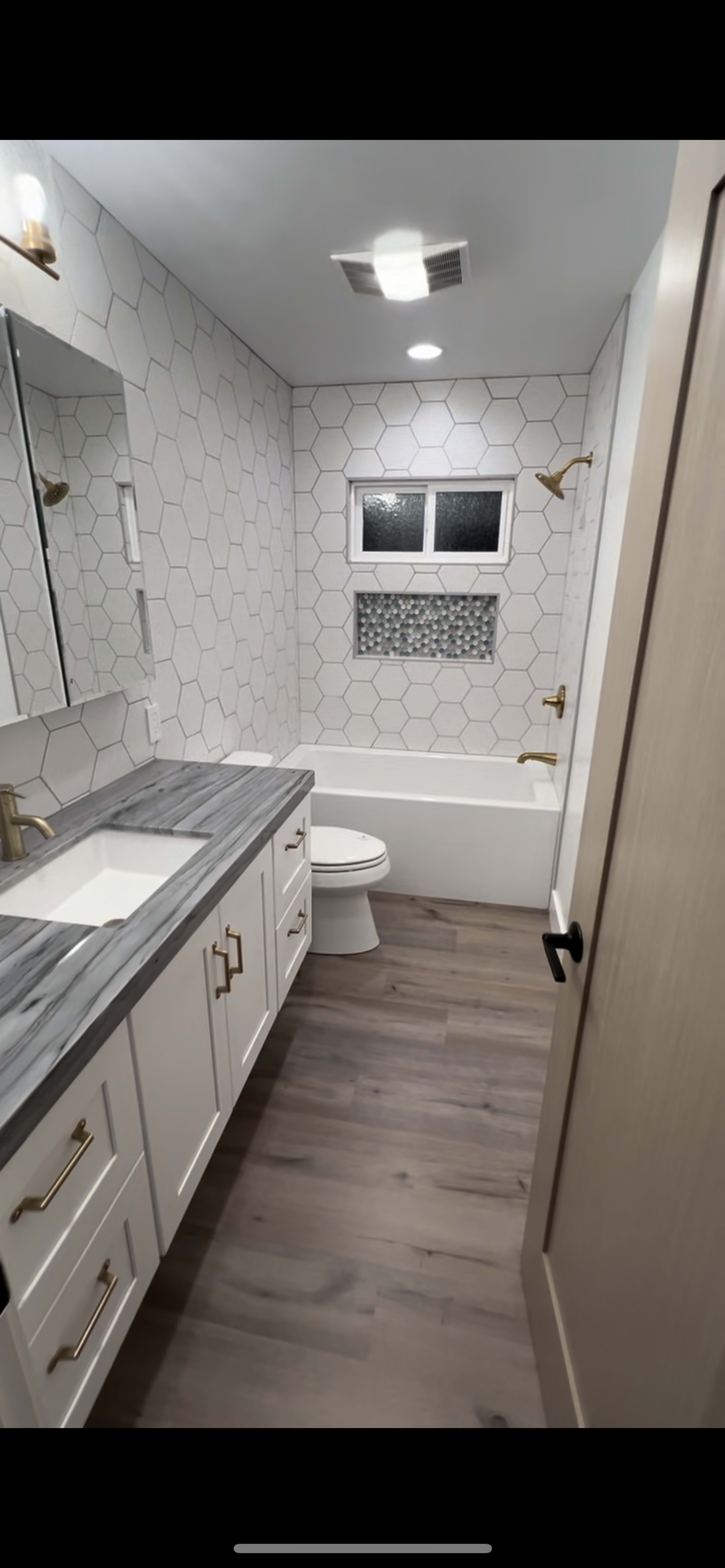 The image shows a modern bathroom featuring a hexagonal tiled wall, a white bathtub, a vanity with a gray countertop, and brass fixtures.