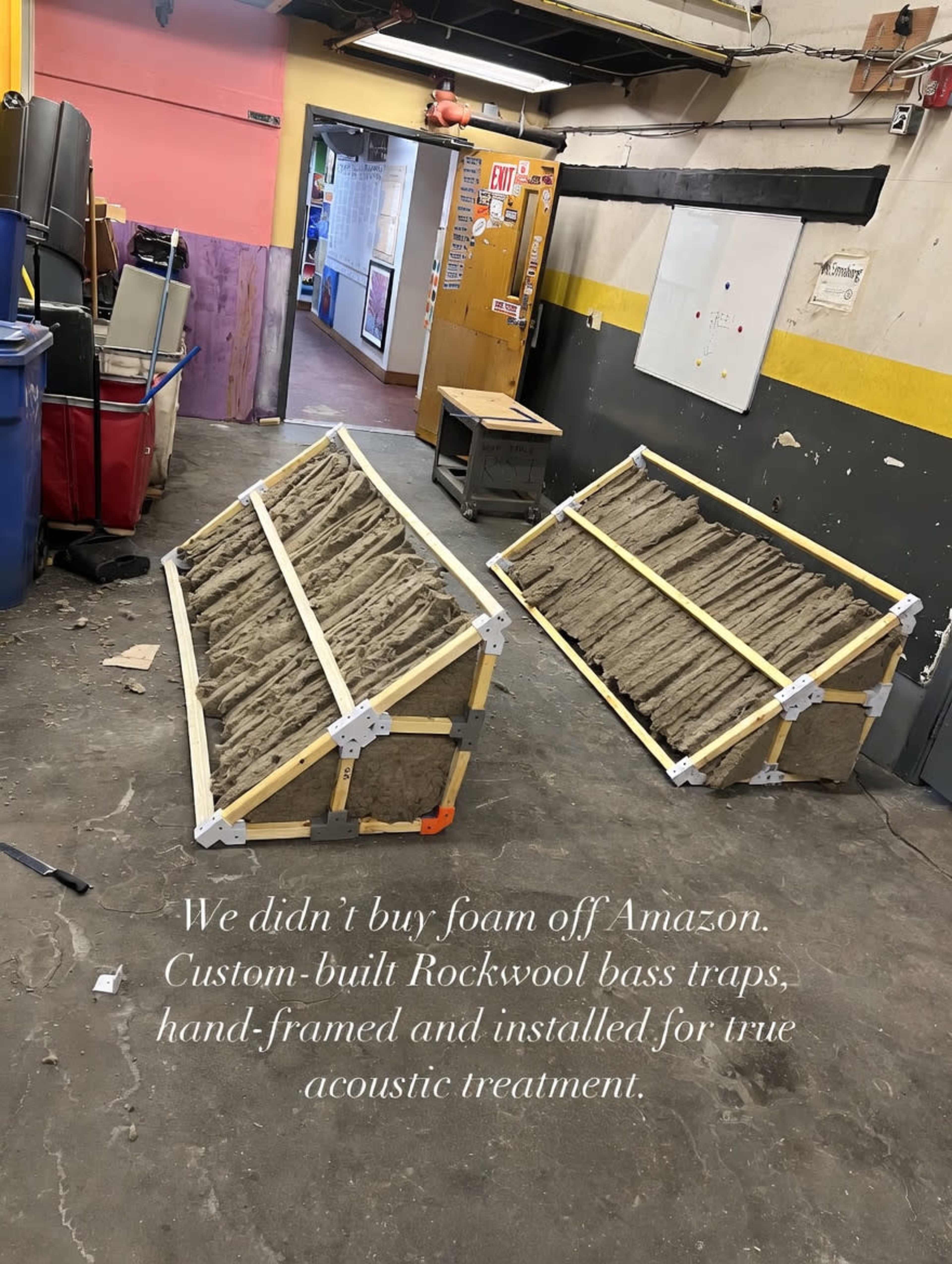 The image shows two custom-built bass traps made of wood and Rockwool, positioned on the floor of a room with colorful walls and various items in the background.