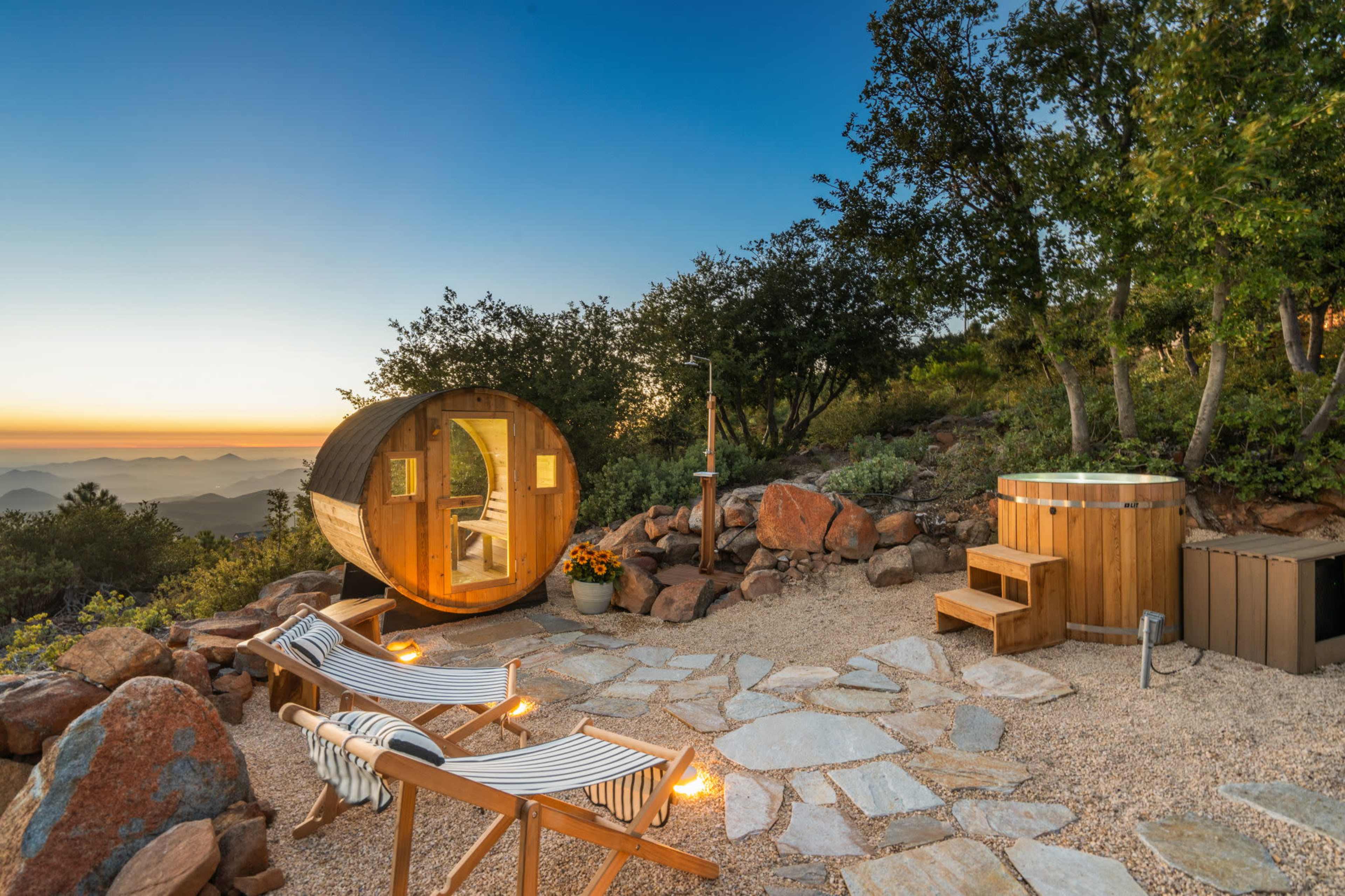 A wooden barrel-shaped cabin sits in a rocky outdoor setting with lounge chairs and a hot tub, surrounded by trees and mountains at sunset.