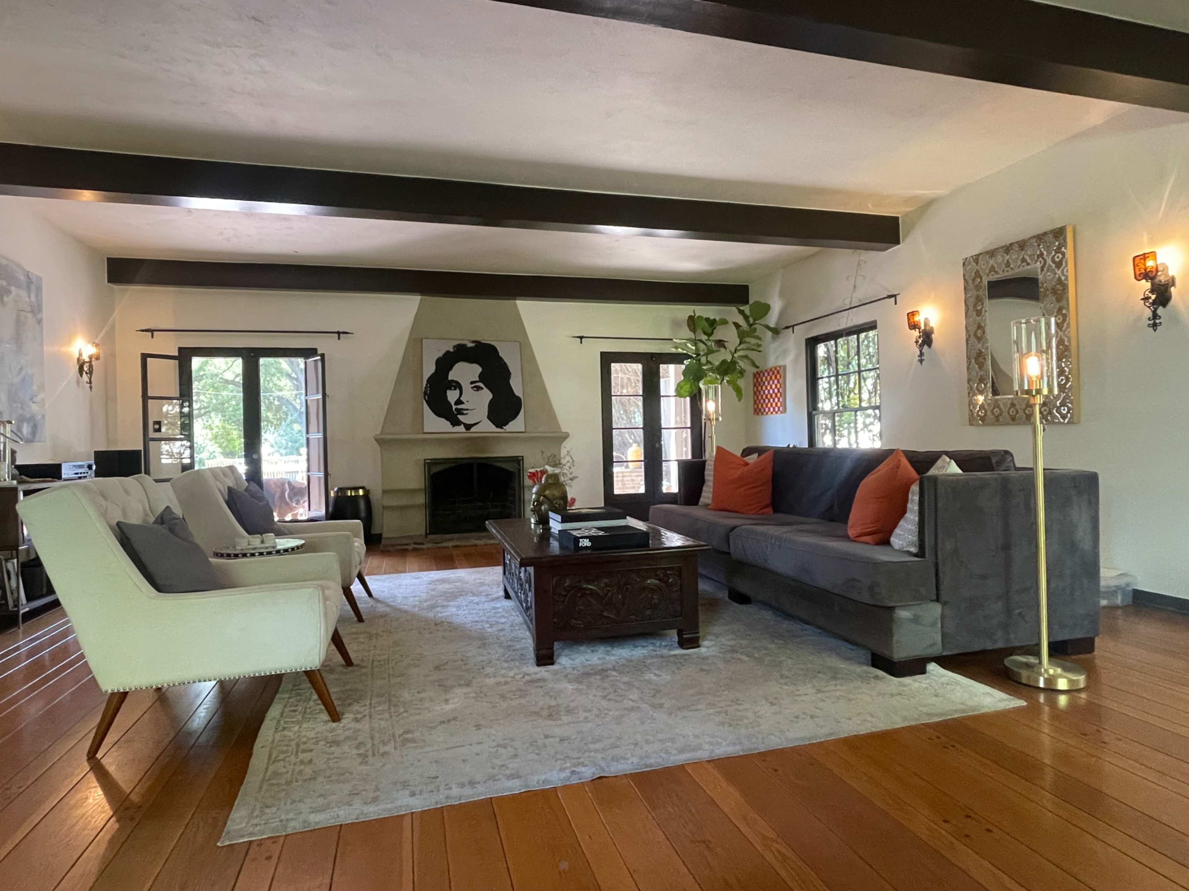 The living room features a gray sectional sofa, two white chairs, a wooden coffee table, and a fireplace, with large windows allowing natural light and a portrait displayed on the wall.