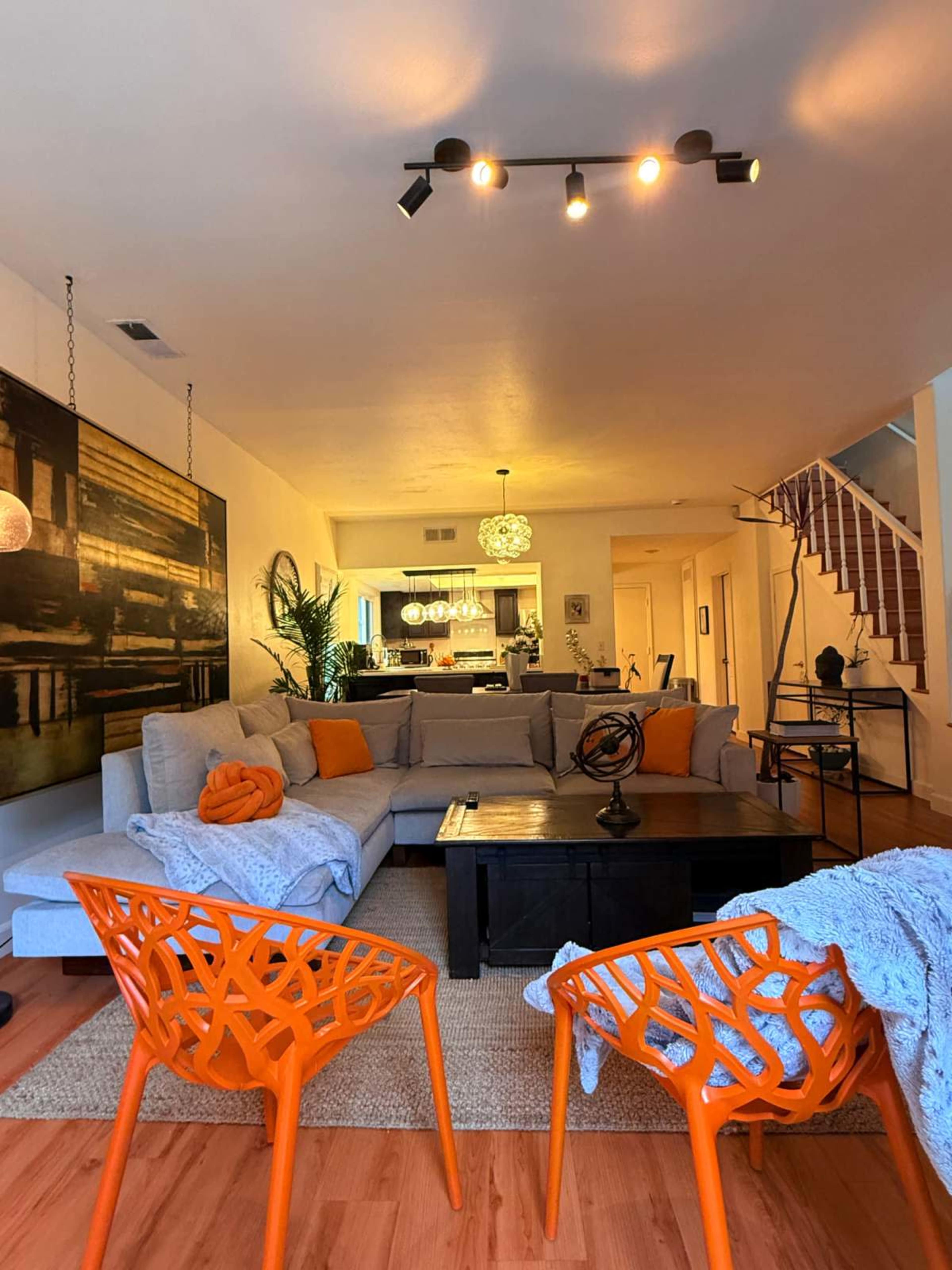The living room features a large gray sectional sofa with orange accent pillows, a dark coffee table, and two bright orange chairs, set against a backdrop of modern decor and an open kitchen.