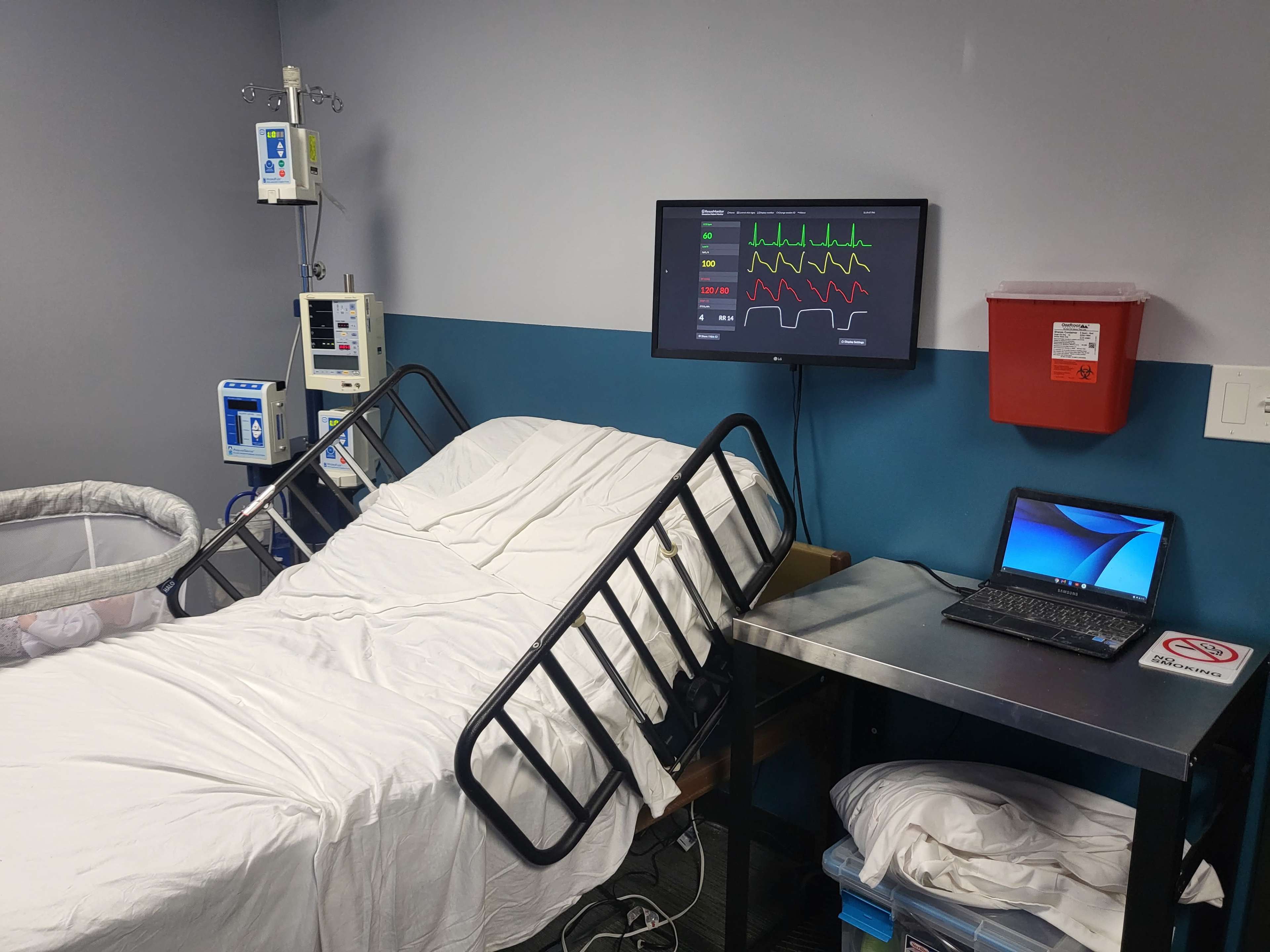 The image shows a hospital room with an unmade bed, monitoring equipment on the wall, and a laptop on a metal table.