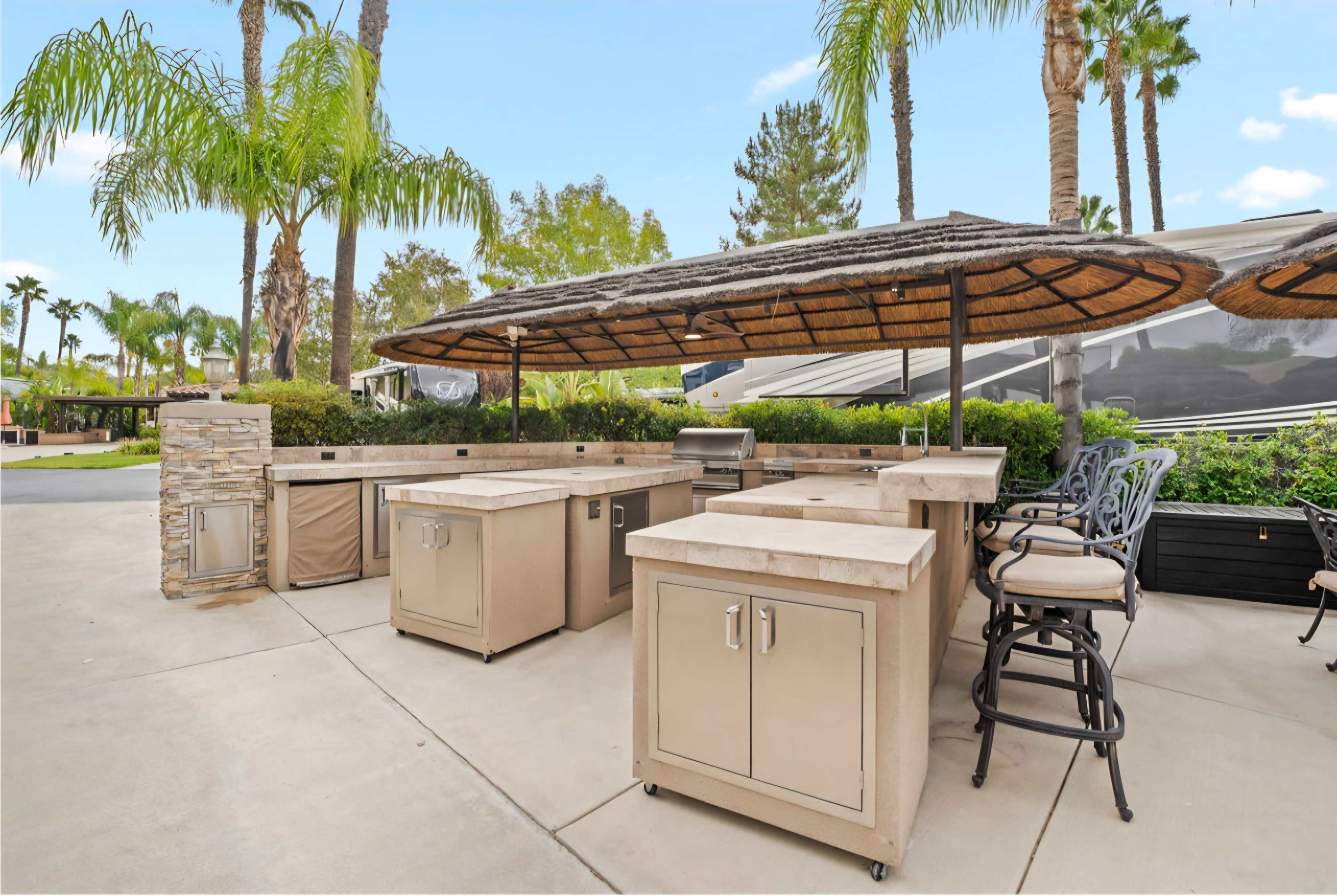 The image shows an outdoor kitchen area with a covered bar, several cabinets, and a grill, surrounded by palm trees and greenery.