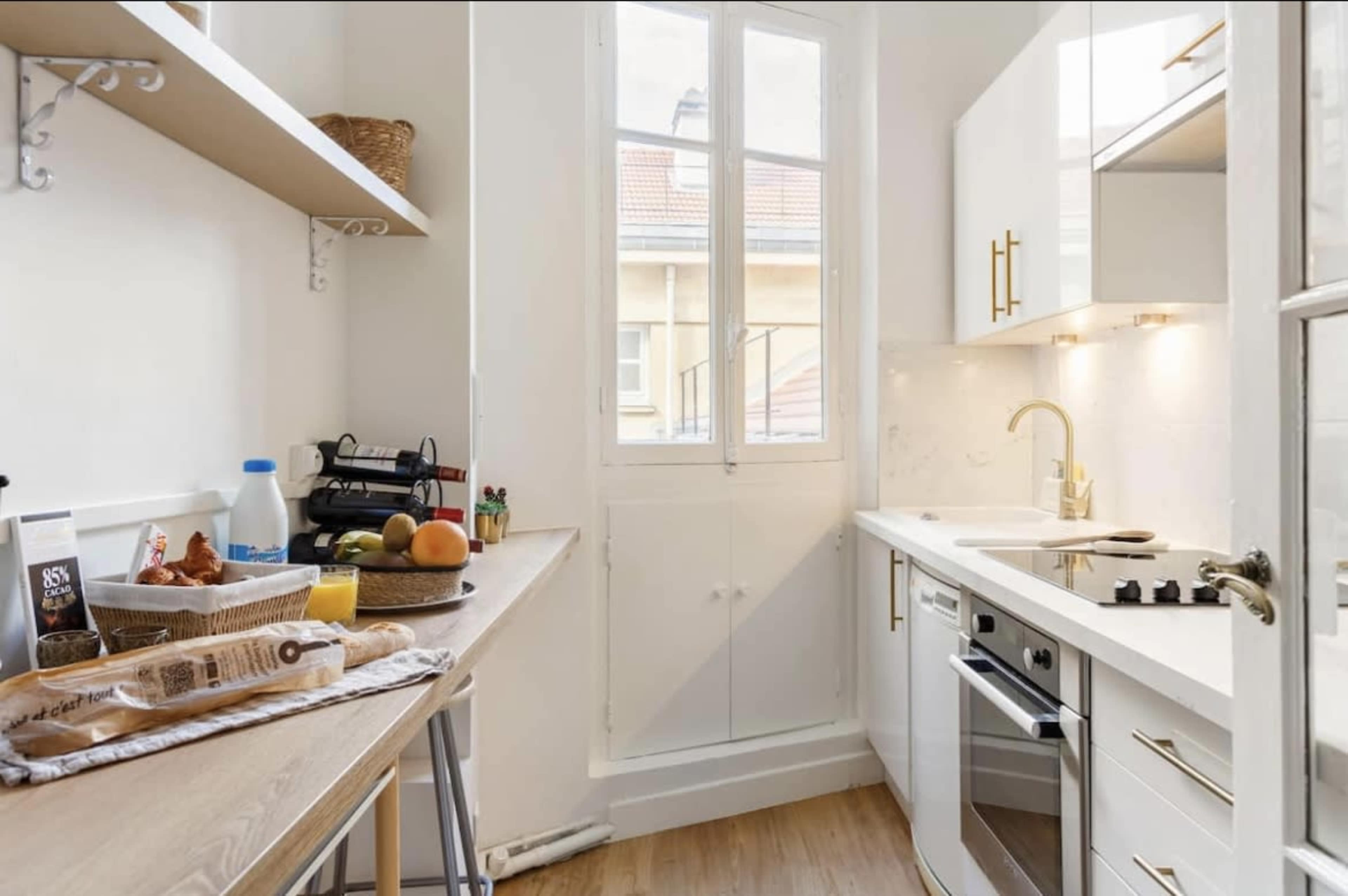 A compact kitchen features white cabinetry, a small dining table with fruits, and a window overlooking an outdoor area.