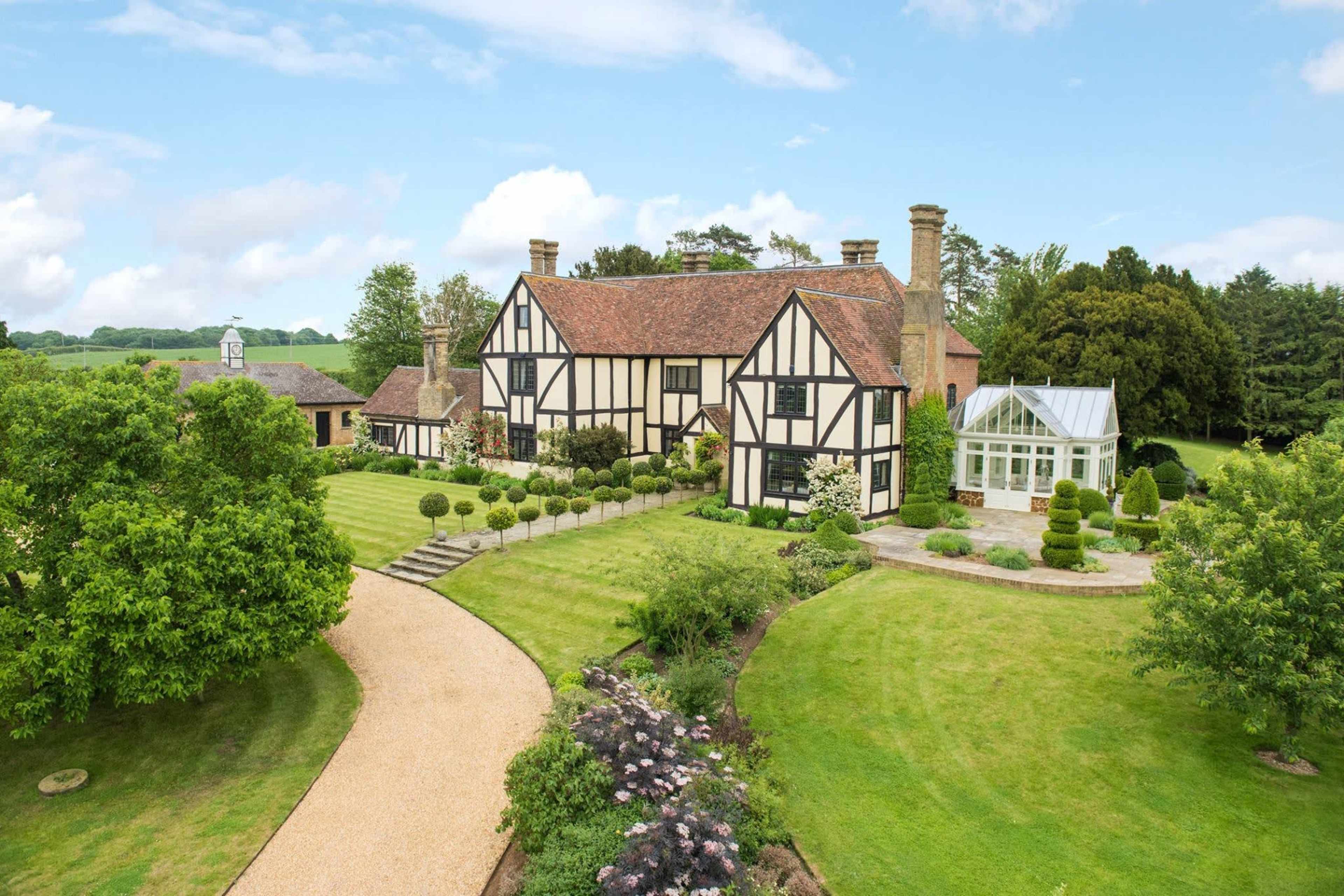 A large, Tudor-style house with a lush lawn and carefully arranged gardens is set against a clear sky.
