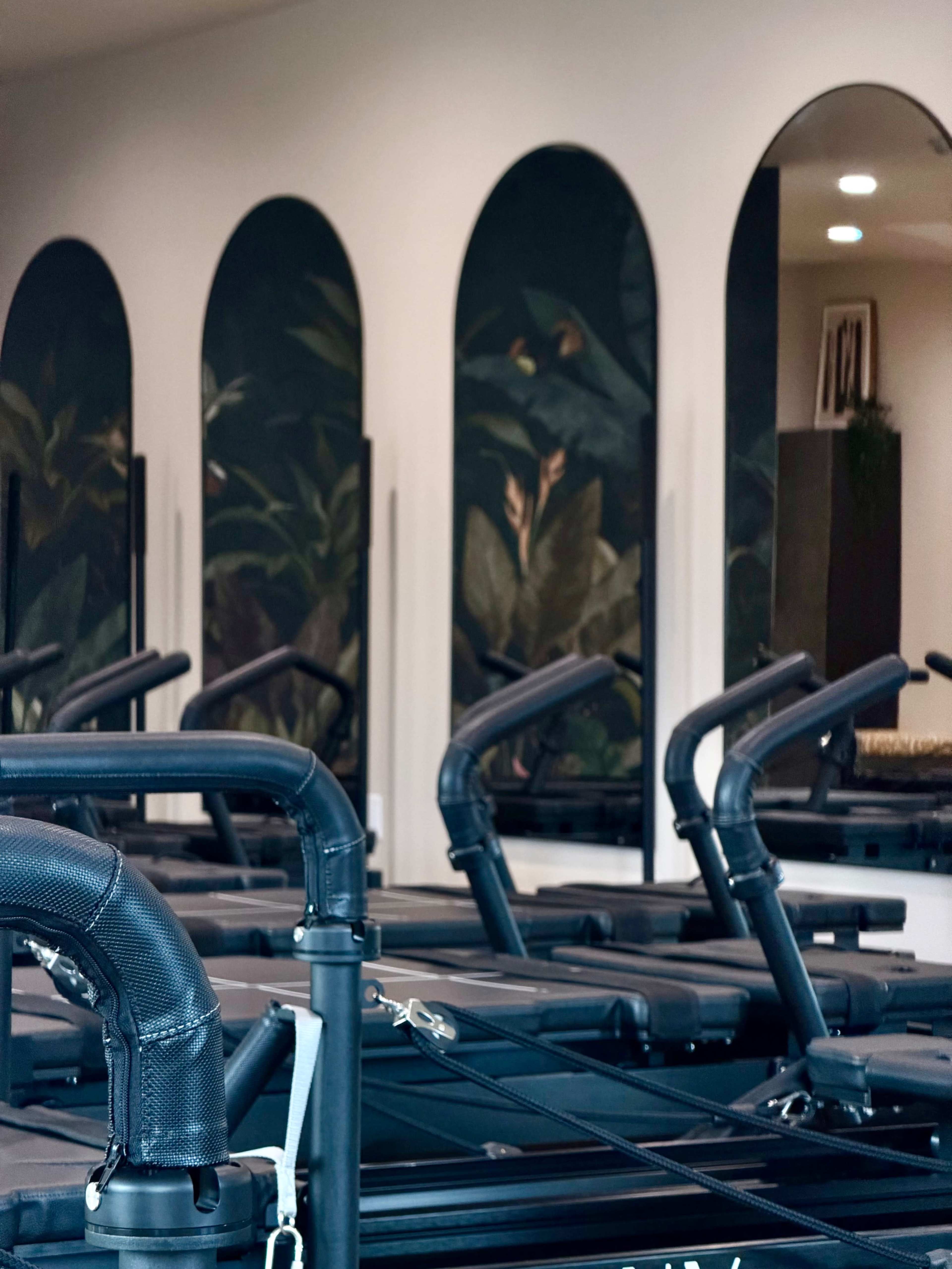 The image shows a modern Pilates studio with several reformer machines and decorative arched mirrors featuring tropical leaf designs on the walls.