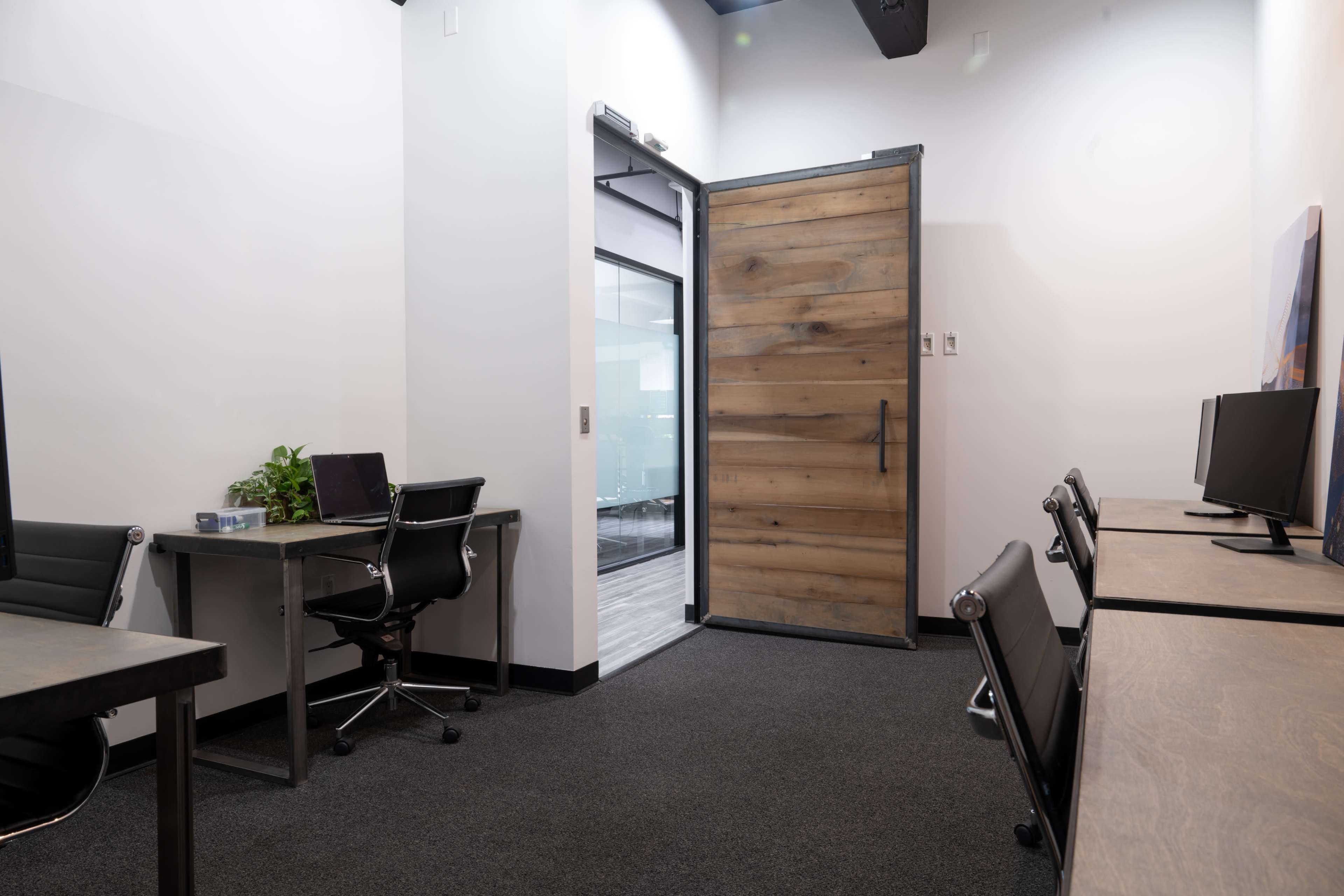 The image shows a modern office space with two desks and chairs facing each other, and a wooden door leading to a glass-enclosed meeting room.