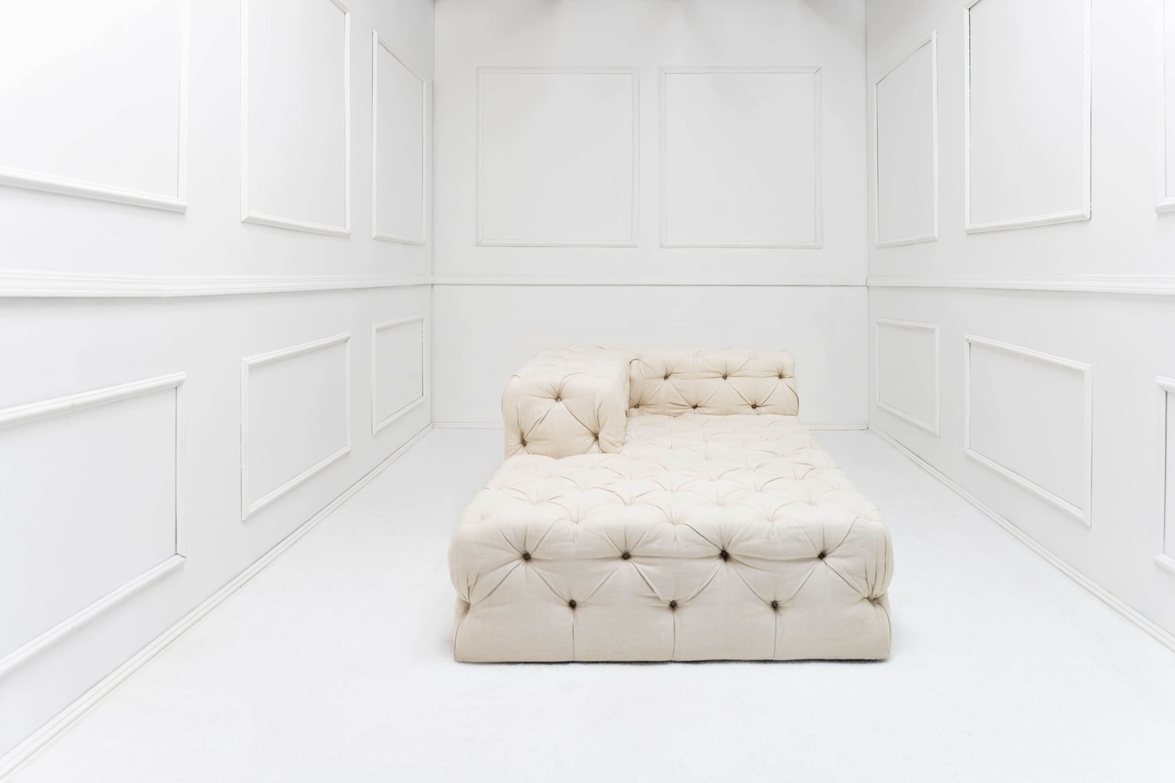 The image shows a minimalist, cream-colored tufted sectional sofa placed in a stark, white room with paneled walls.