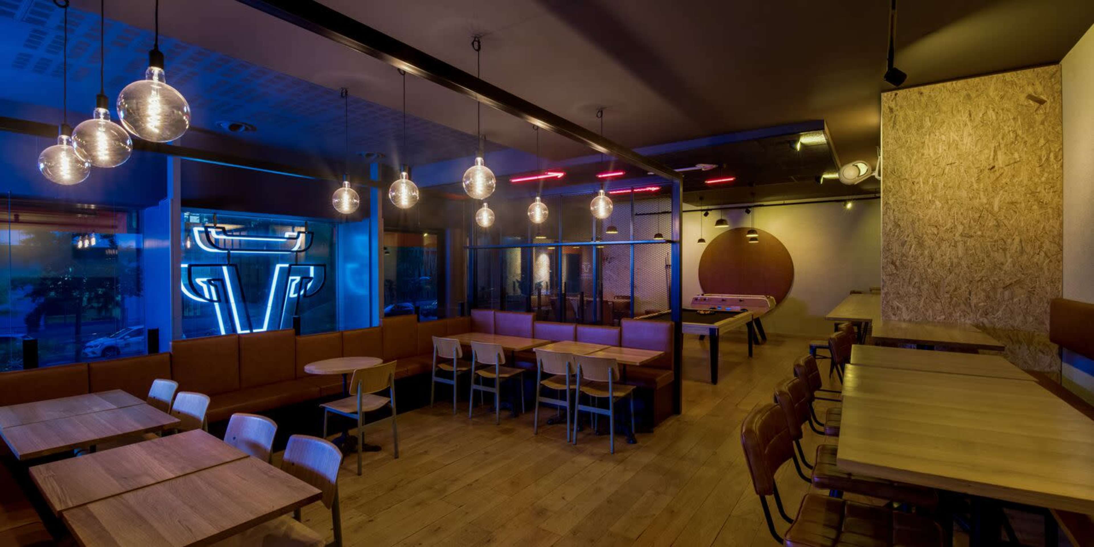 A modern cafe interior with wooden tables and benches, illuminated by pendant lights, featuring a neon sign and a game table in the background.