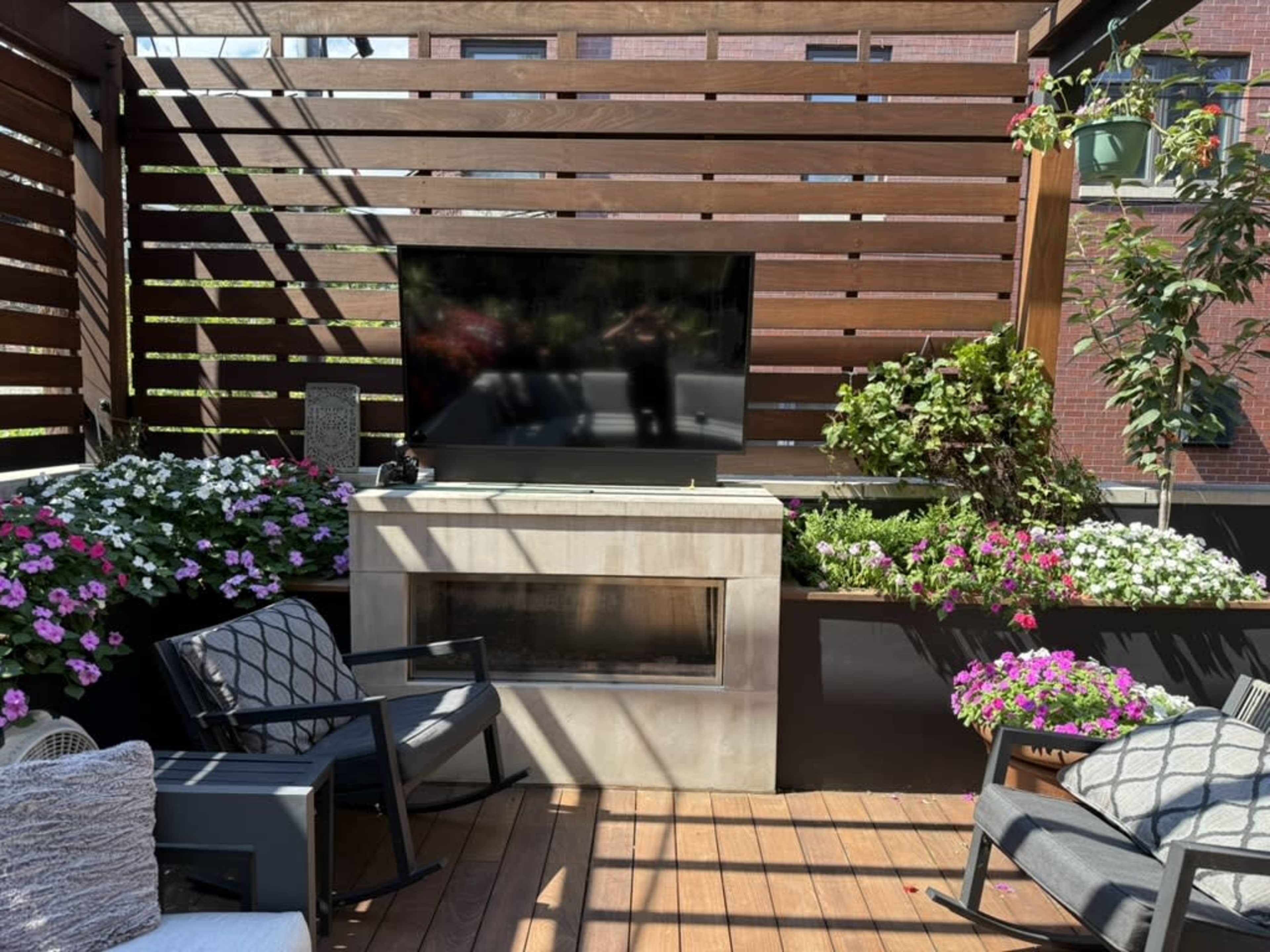 The image shows a patio area featuring a mounted television, a gas fireplace, and planters filled with colorful flowers, surrounded by wooden slats for privacy.