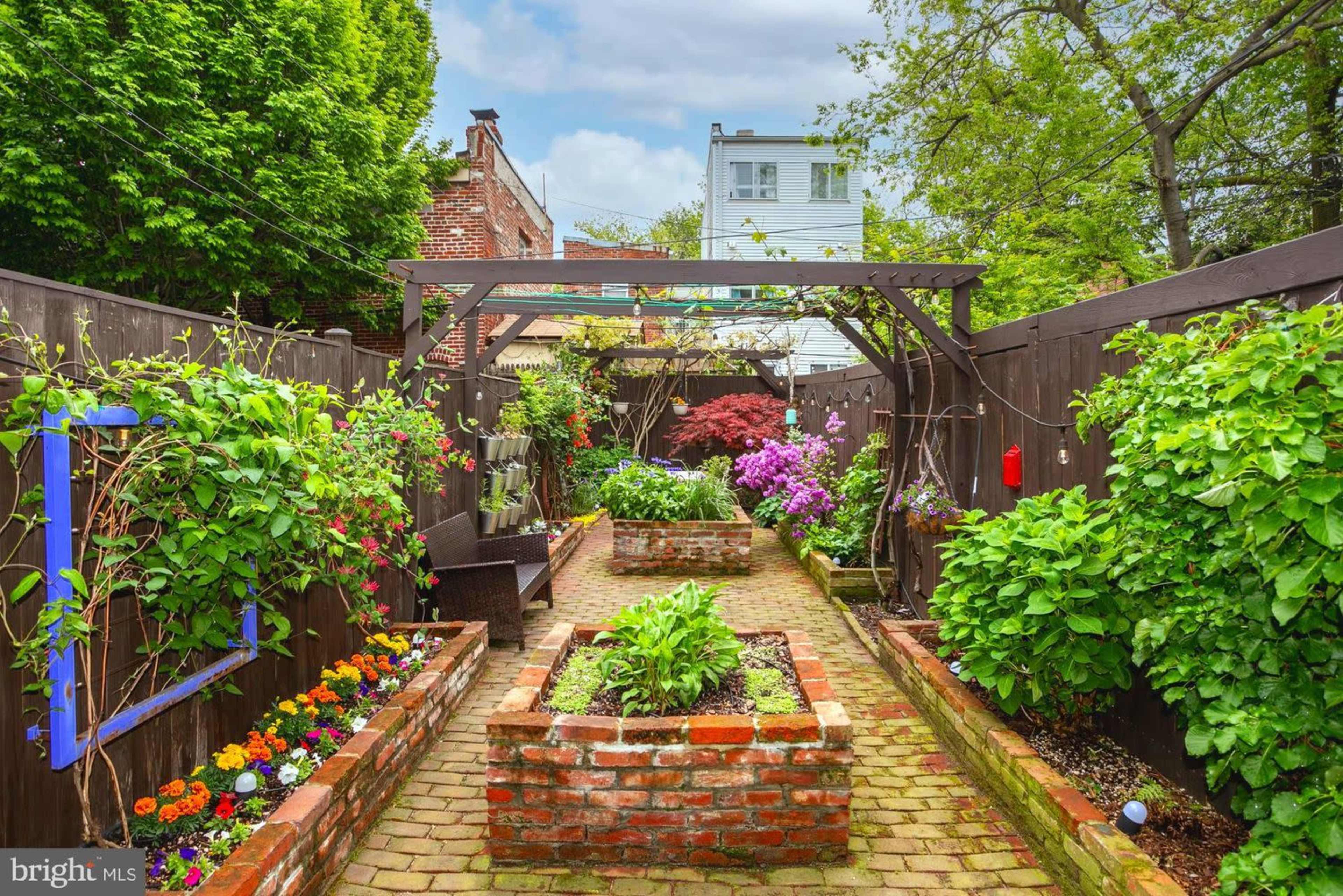 The image shows a landscaped garden pathway bordered by raised brick planters filled with vibrant flowers and greenery, set against wooden fencing.