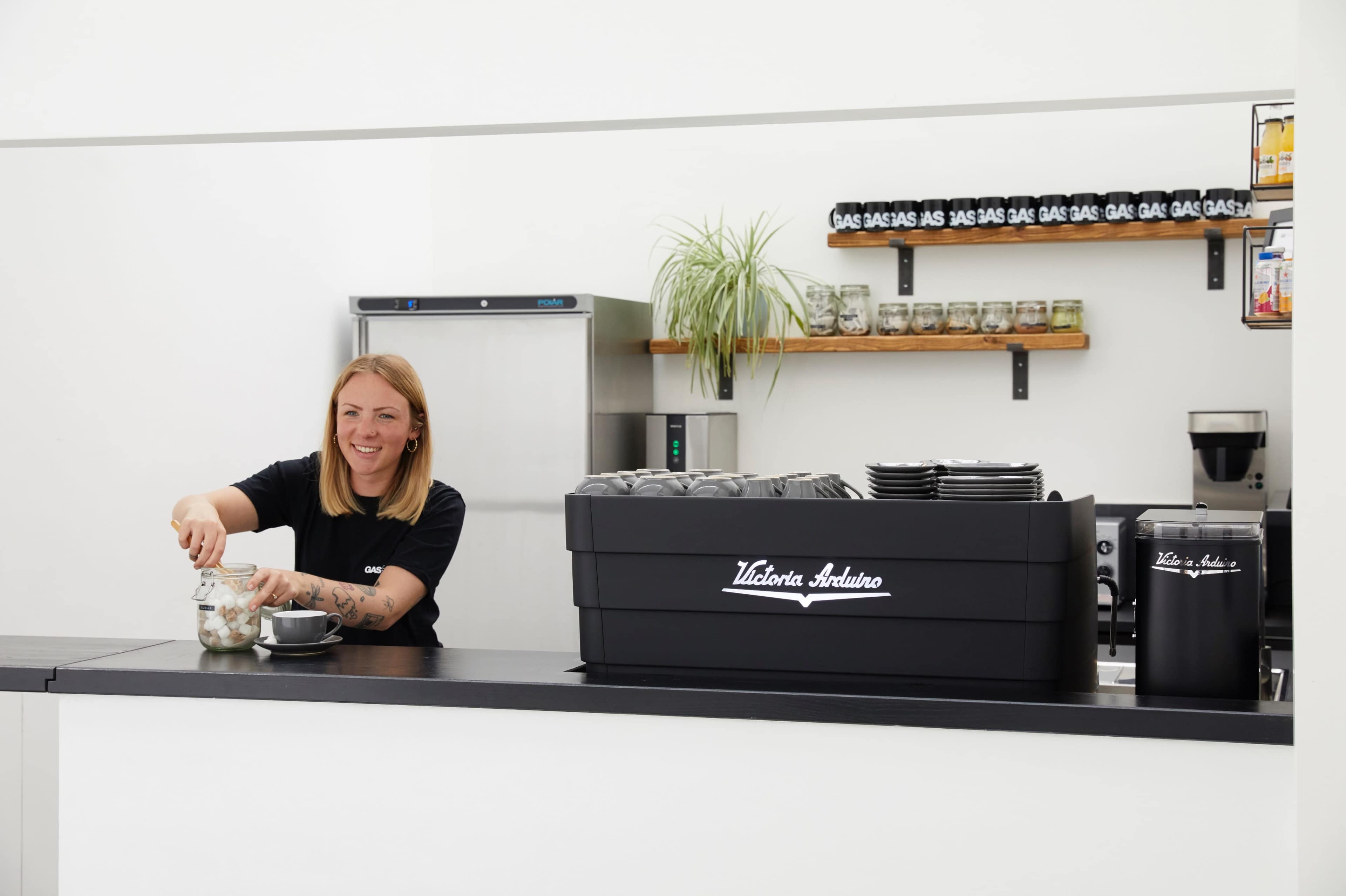 A barista at a café serves a drink while standing behind a sleek counter with coffee equipment and jars displayed.