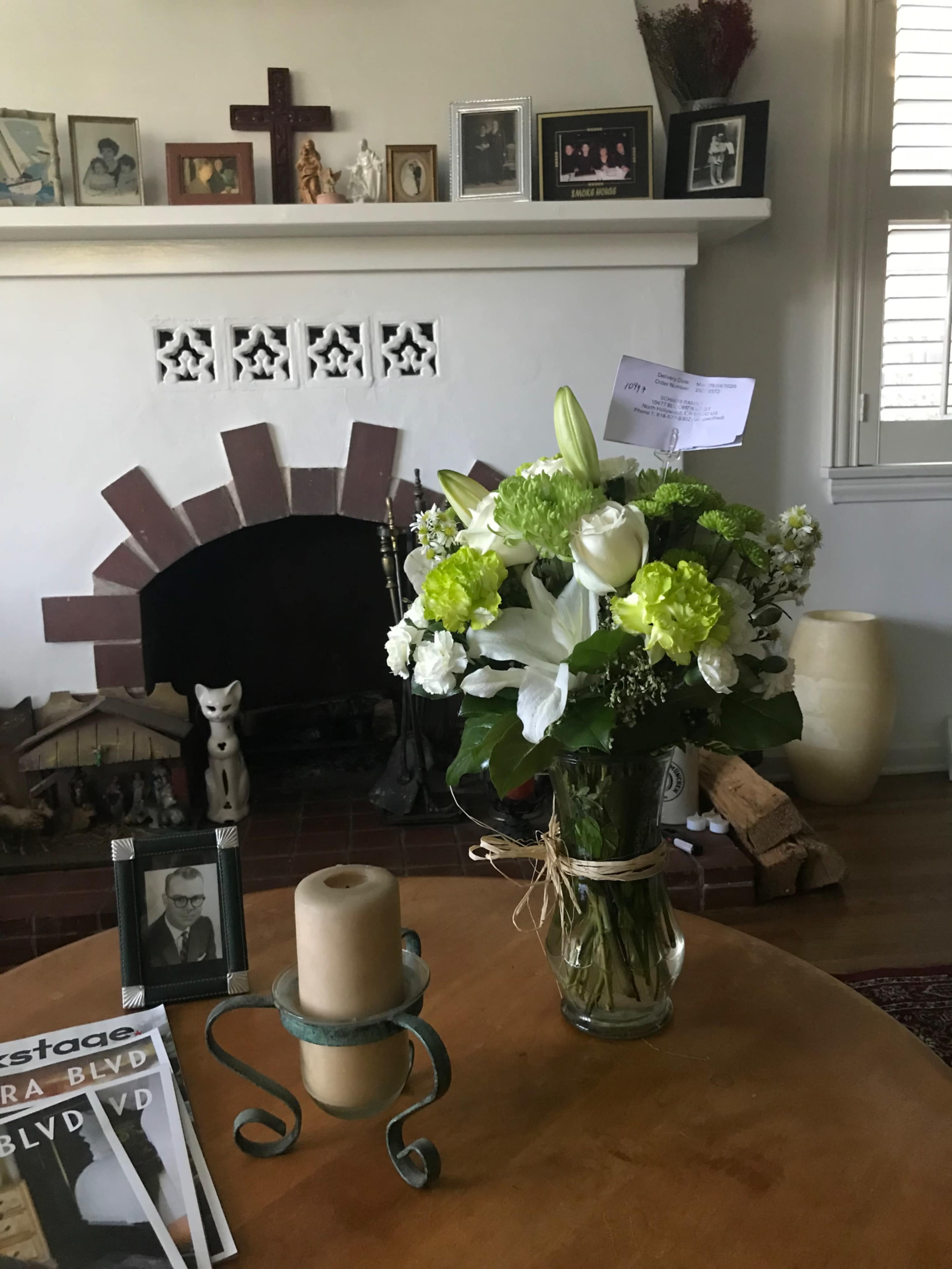 A vase of flowers sits on a wooden table in a living room that features a fireplace and various framed photos on the wall.