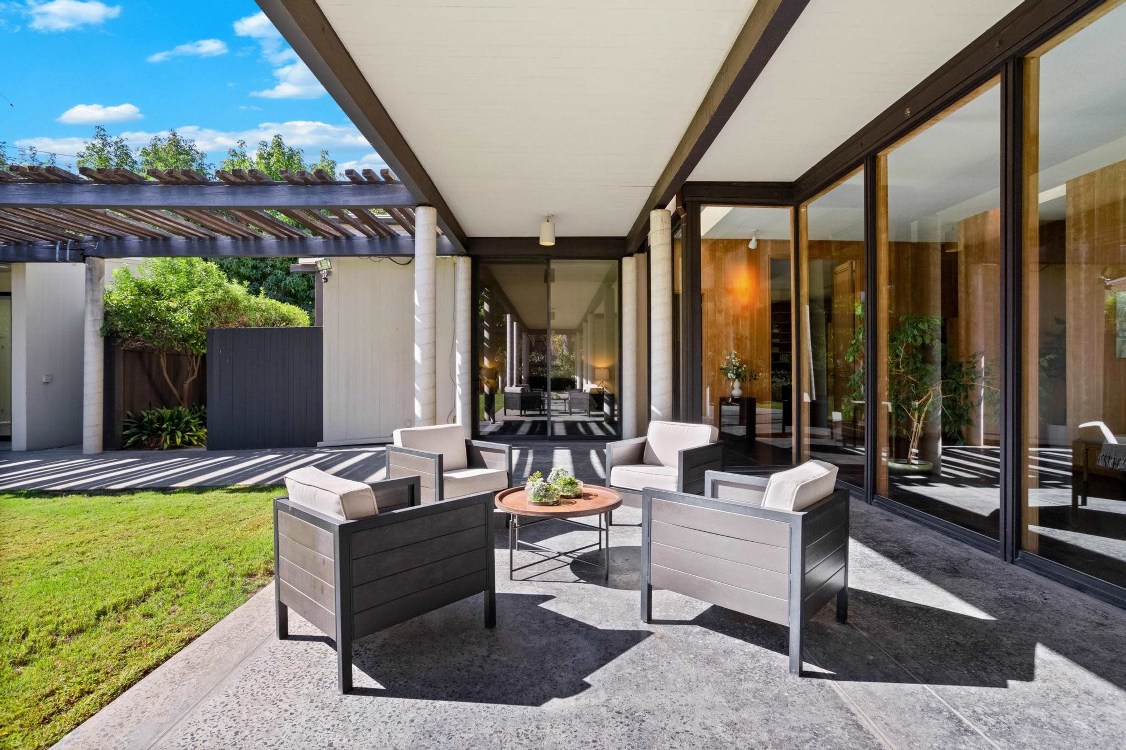 A seating area with four chairs and a small table is positioned on a patio next to large glass doors, surrounded by greenery.