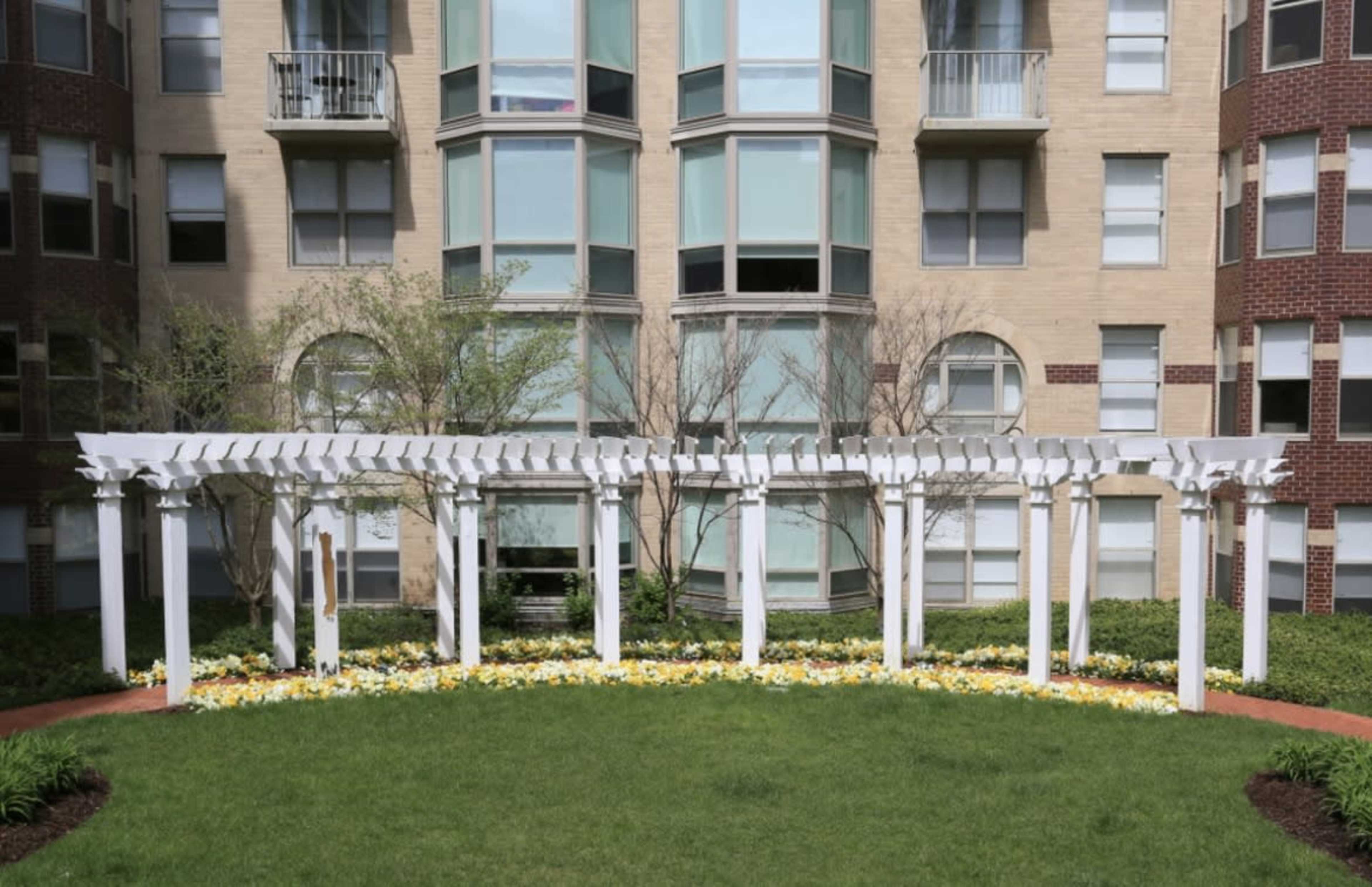 The image shows a landscaped area featuring a white pergola surrounded by flower beds and grass, in front of an apartment building with large windows.