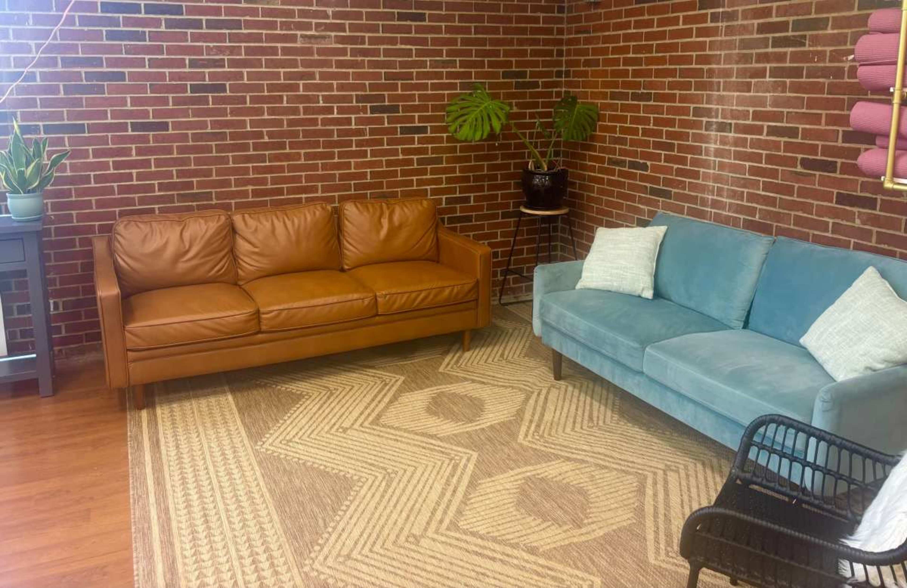 The image shows a cozy living space with a brown leather sofa and a light blue fabric couch, positioned on a geometric-patterned rug against a backdrop of exposed brick walls.