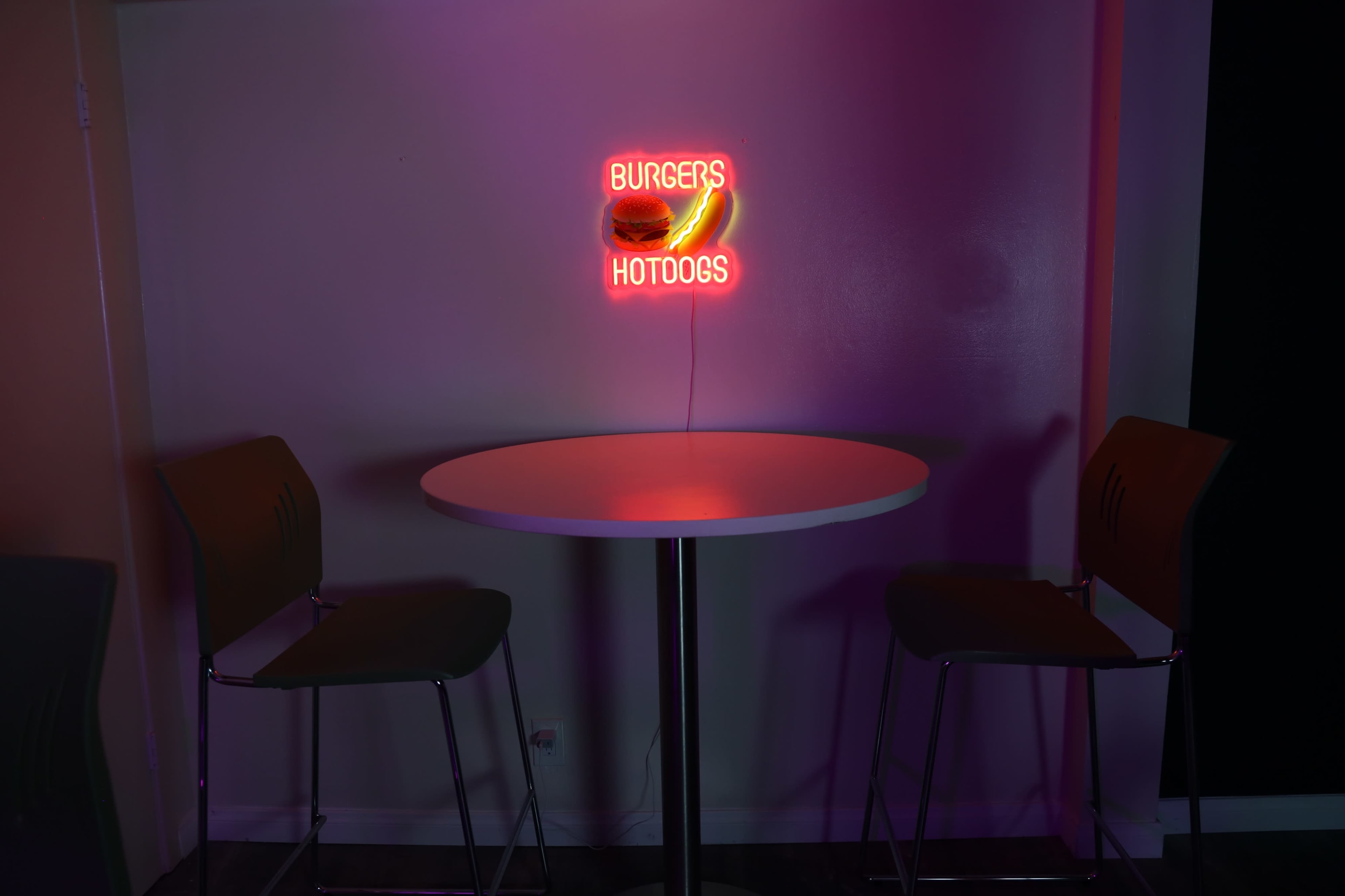 A small table with two stools is illuminated by a neon sign that reads "BURGERS" and "HOTDOGS."