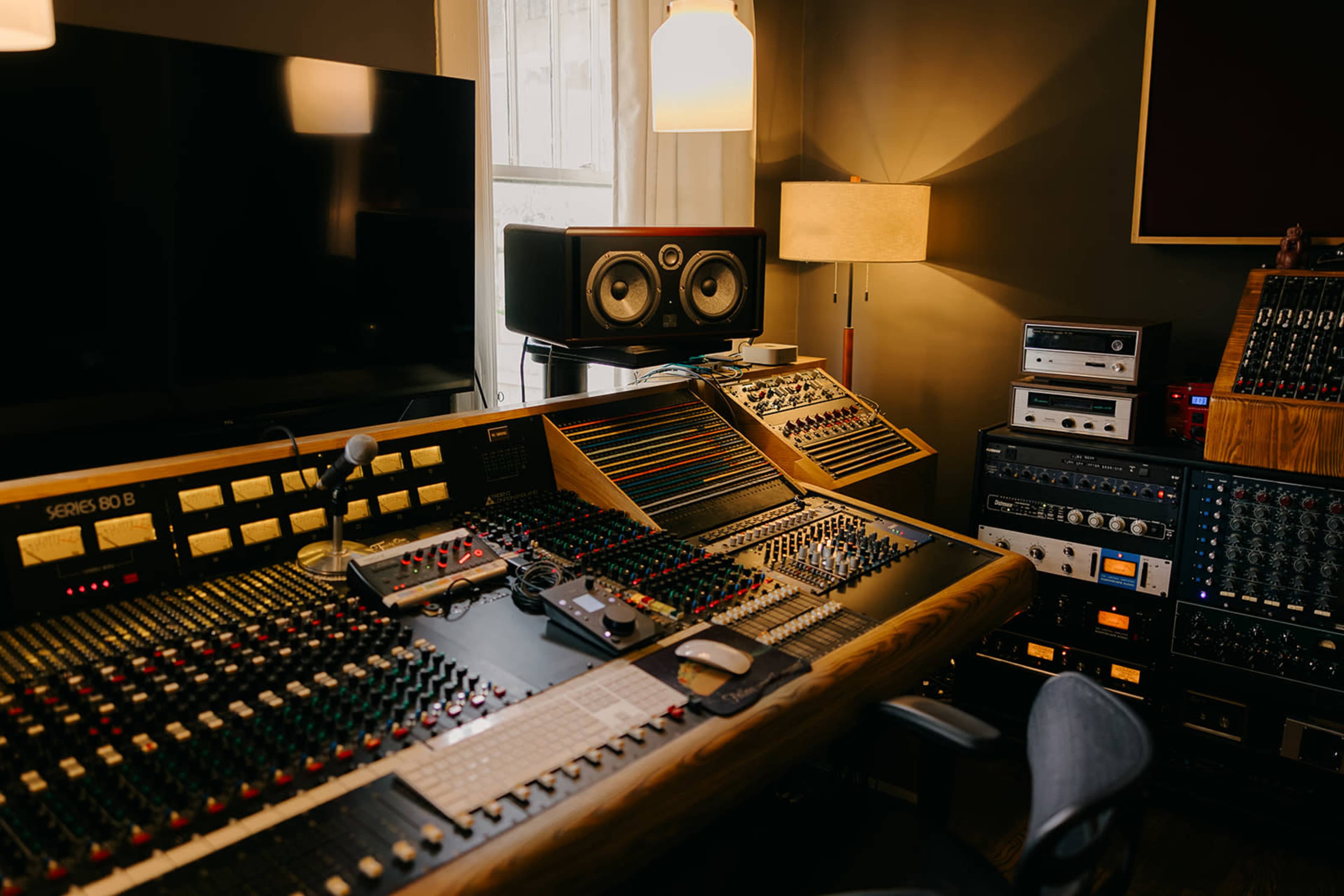 The image shows a sound studio control room featuring a large mixing console with various knobs and sliders, studio monitors, and audio equipment arranged on shelves.