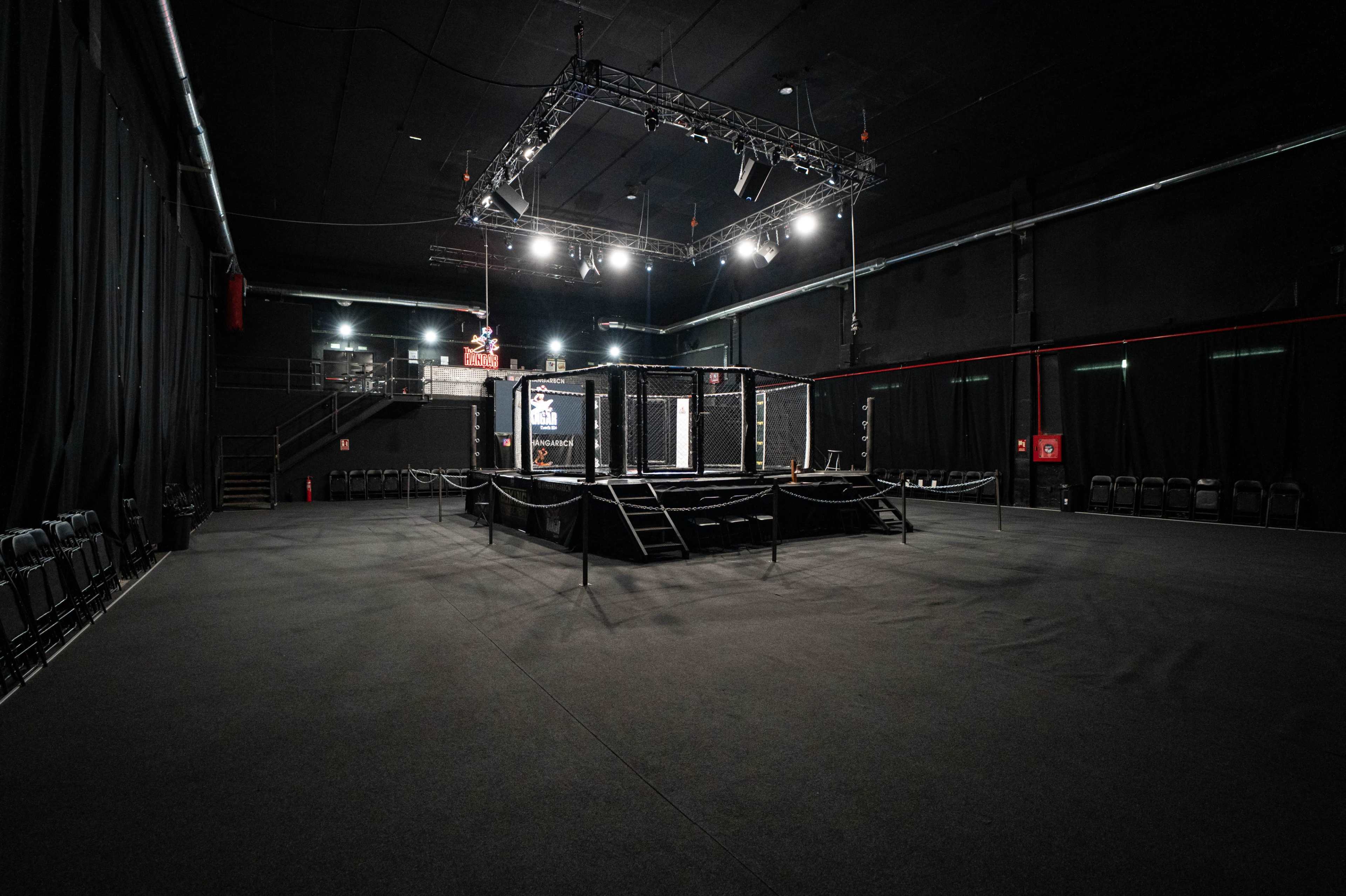 The image shows a dimly lit indoor arena featuring an octagonal fighting cage at the center, surrounded by rows of empty chairs and a large spotlight above.