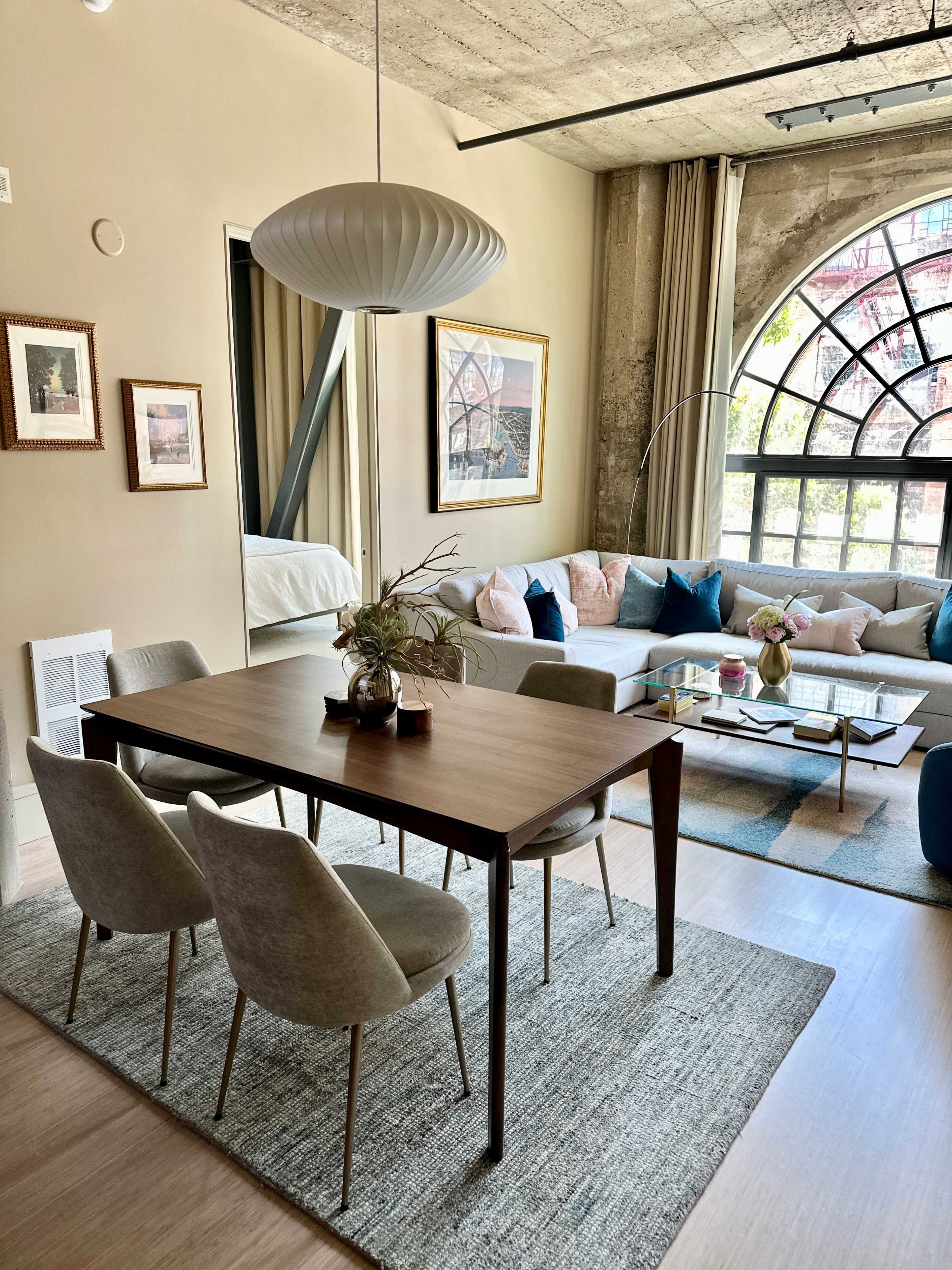 A modern living room with a dining table and chairs, a large window featuring an arched design, and a combination of furniture and decor in neutral tones.