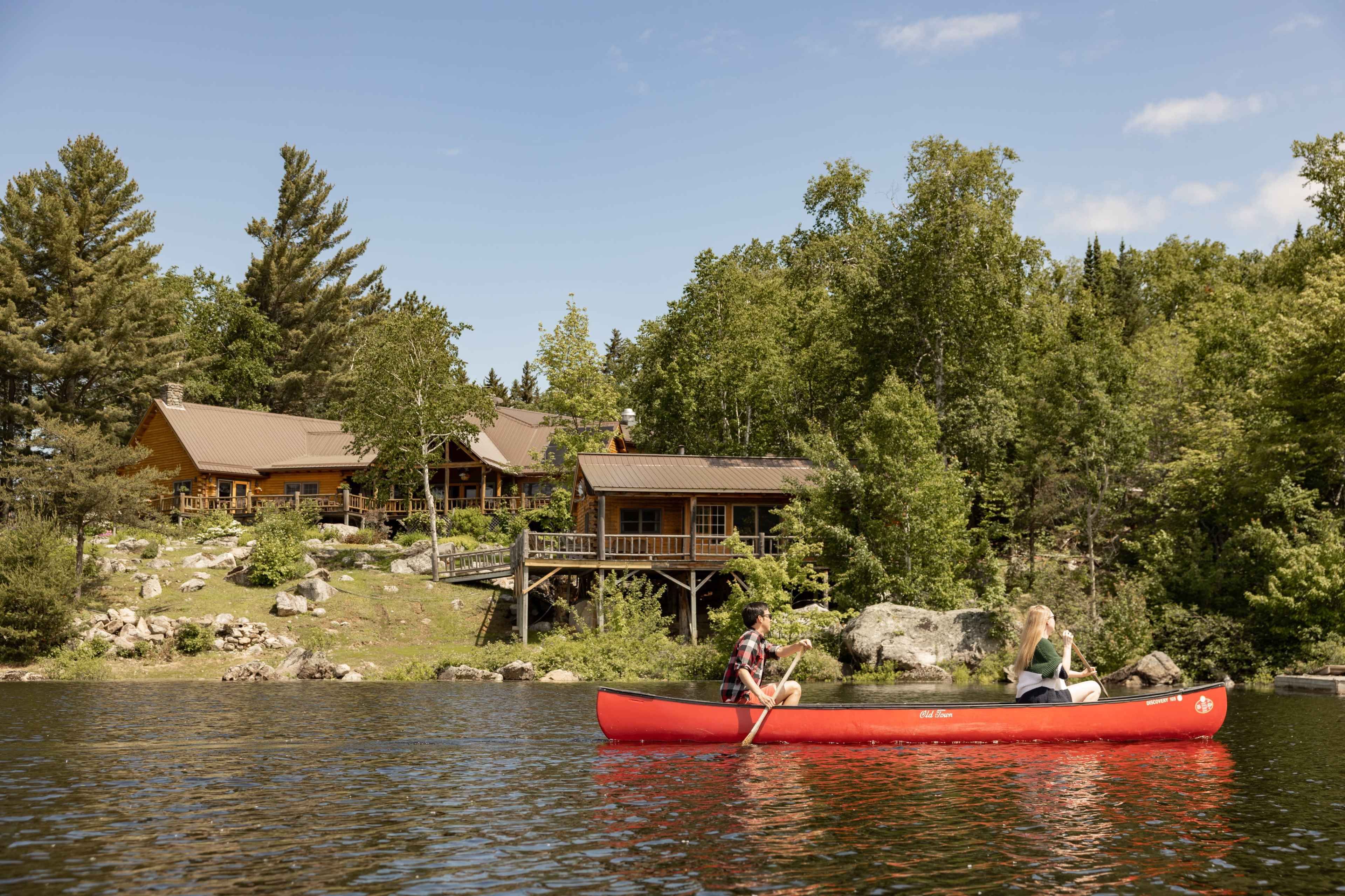 Two people are paddling a red canoe on a calm lake in front of a wooden cabin surrounded by trees.