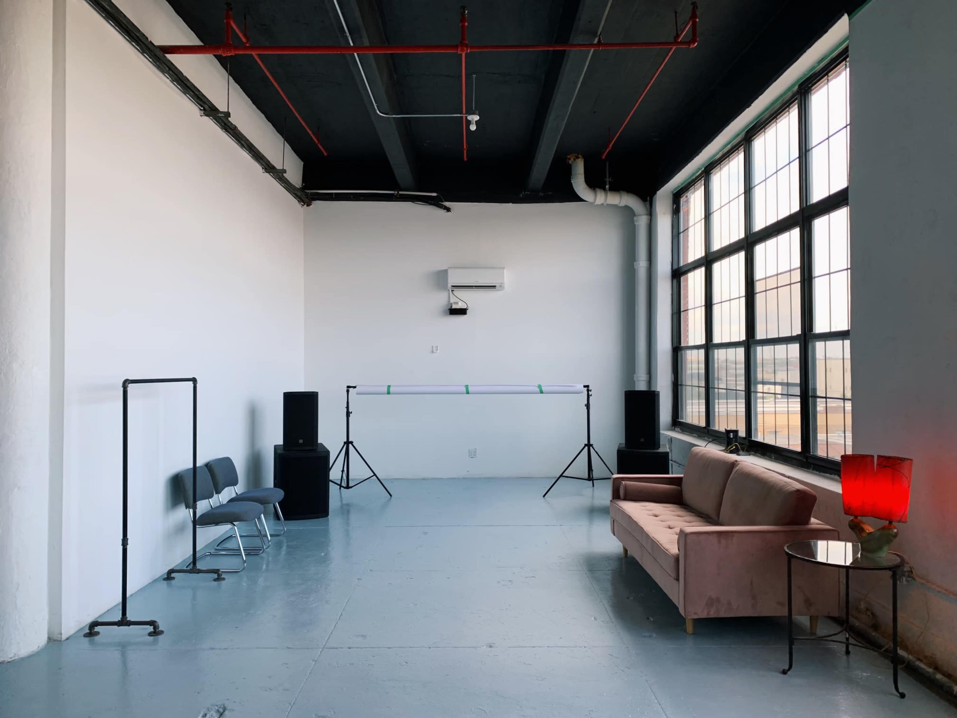 A minimalistic indoor space with a pink sofa, a side table with a red lamp, and audio equipment positioned beside a large window.
