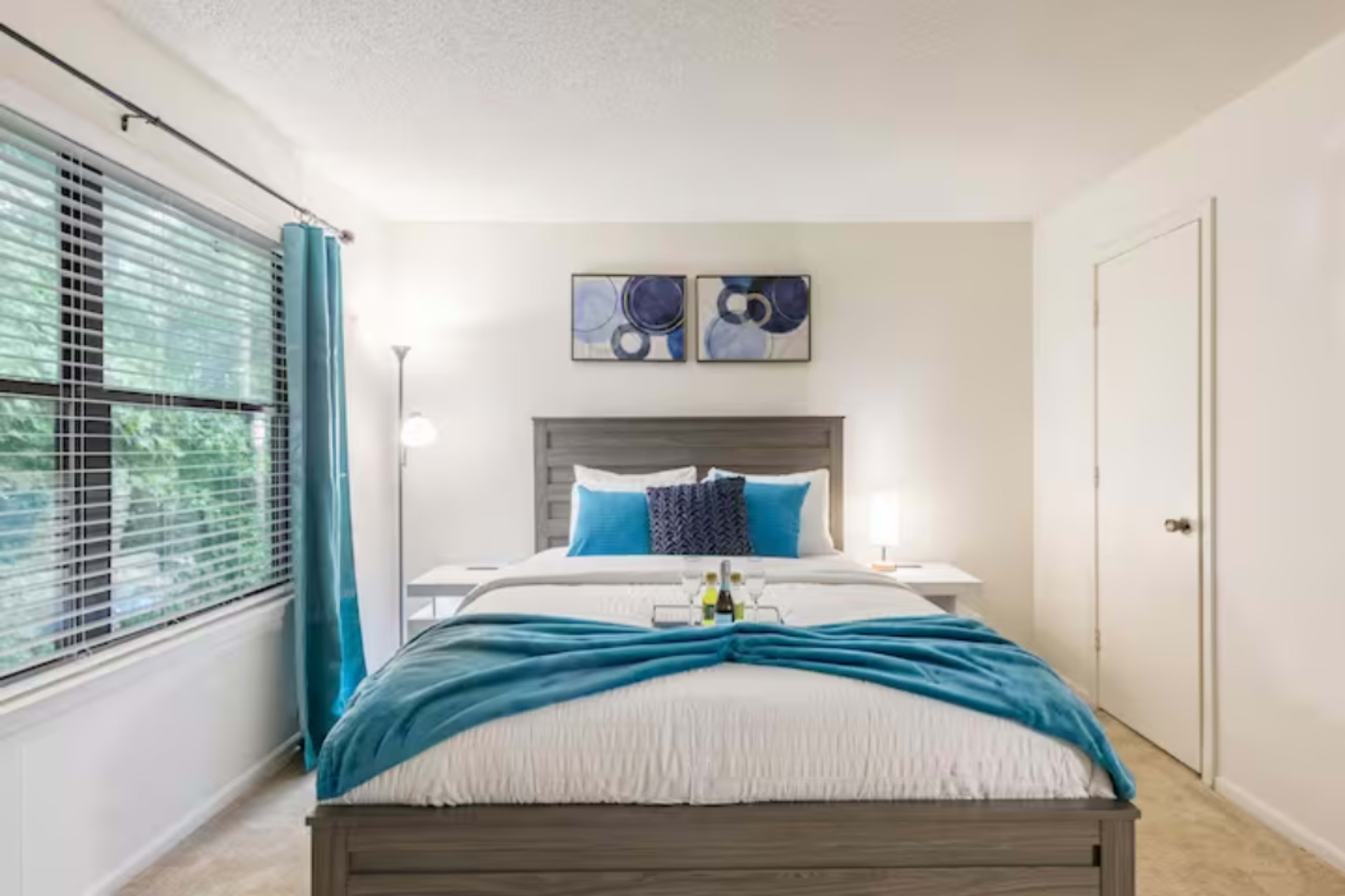 A bedroom with a large bed adorned with a blue throw blanket, two bedside tables with lamps, and a window featuring sheer curtains.