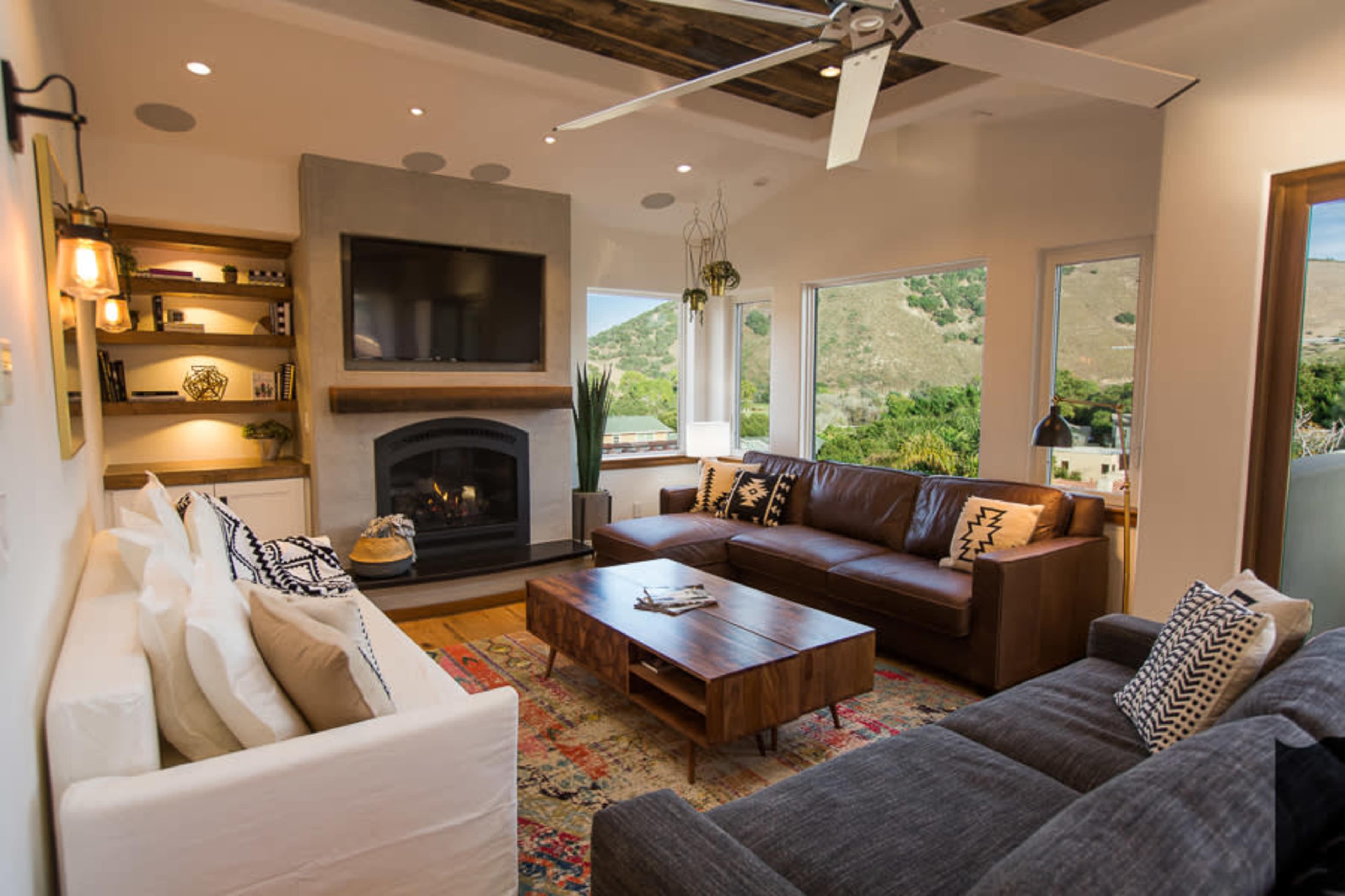 The living room features a large sectional sofa, a wooden coffee table, a cozy fireplace, and a mounted television, with windows offering a view of the hillside.