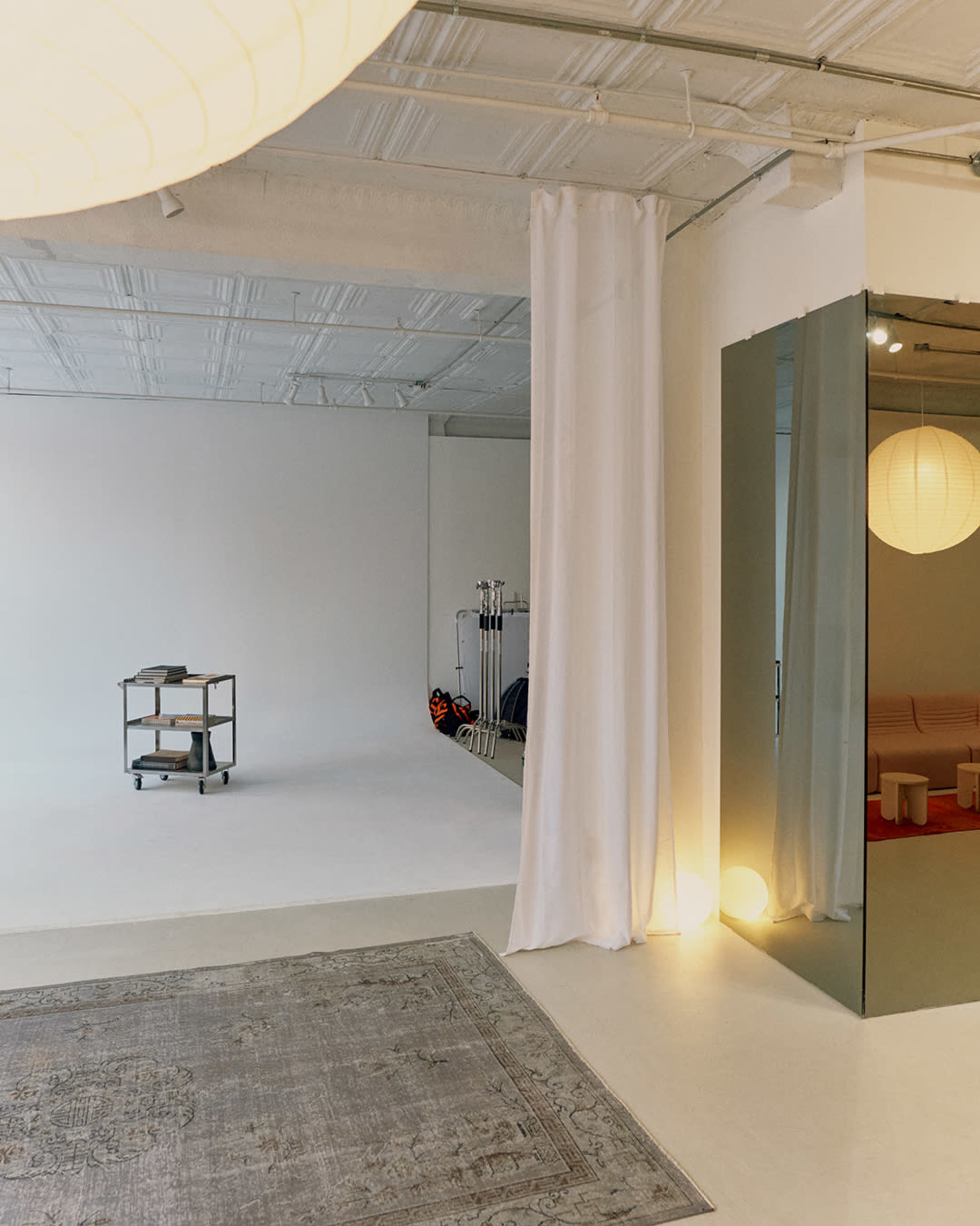 The image shows a spacious, minimalistic room with a blank white backdrop, a rolling cart, soft lighting, and a large mirror reflecting a portion of the space.