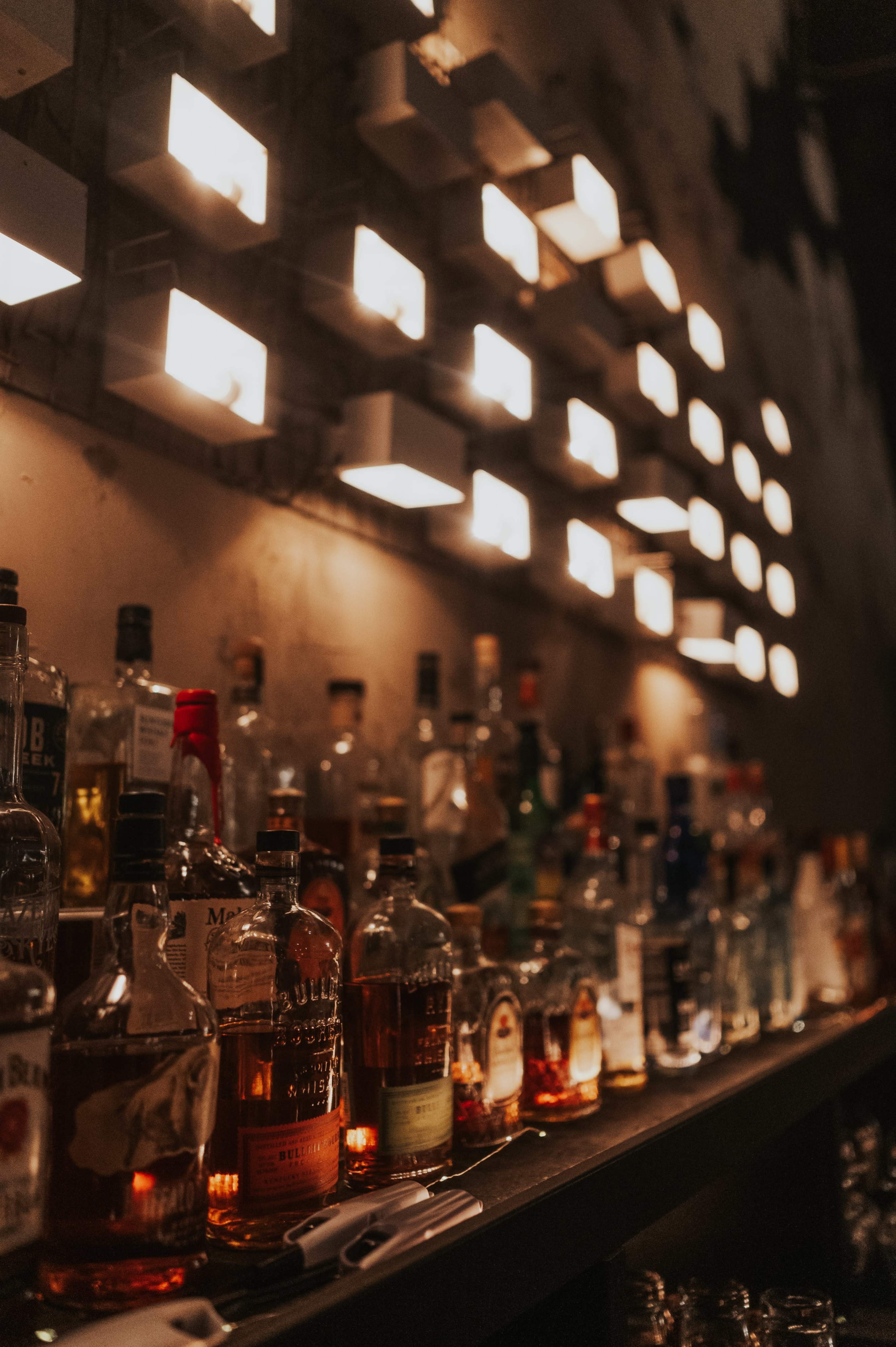 The image shows a bar shelf lined with various bottles of liquor illuminated by wall-mounted lights.