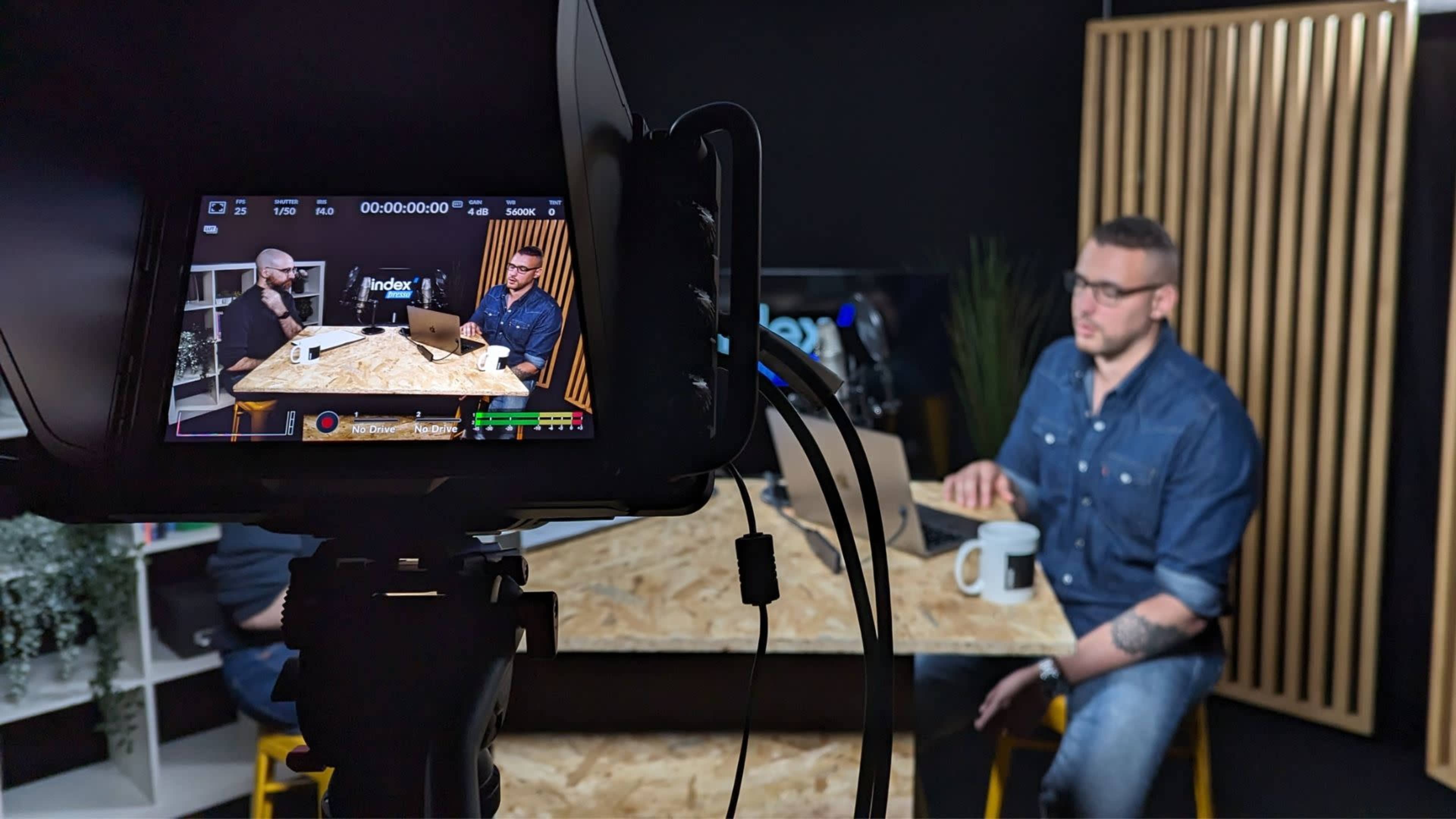 A video recording setup with two people engaged in a discussion at a wooden table, viewed through a camera lens.