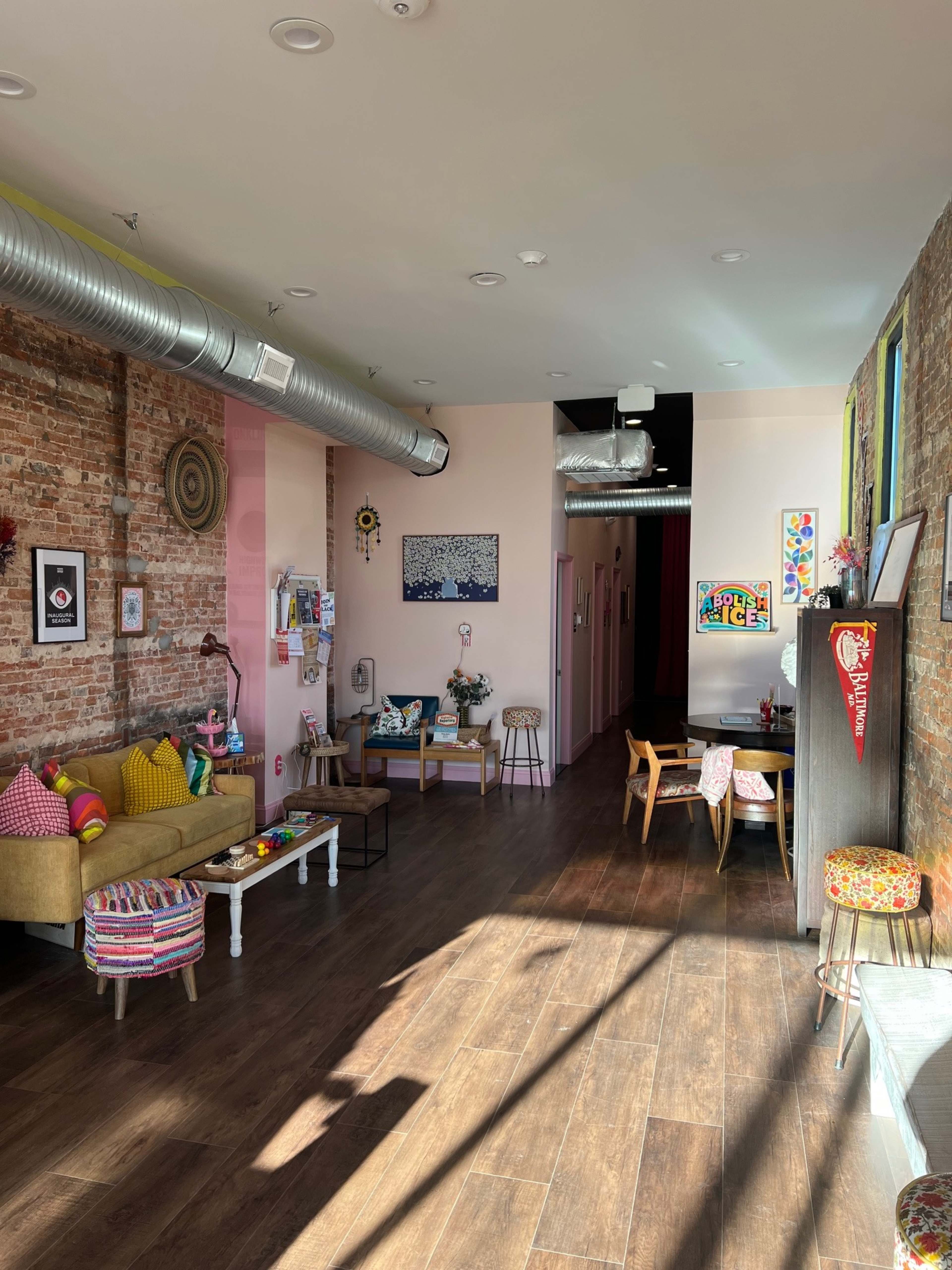 The image shows a brightly lit interior space featuring a cozy seating area with a couch, a small dining table, and artwork on the walls.