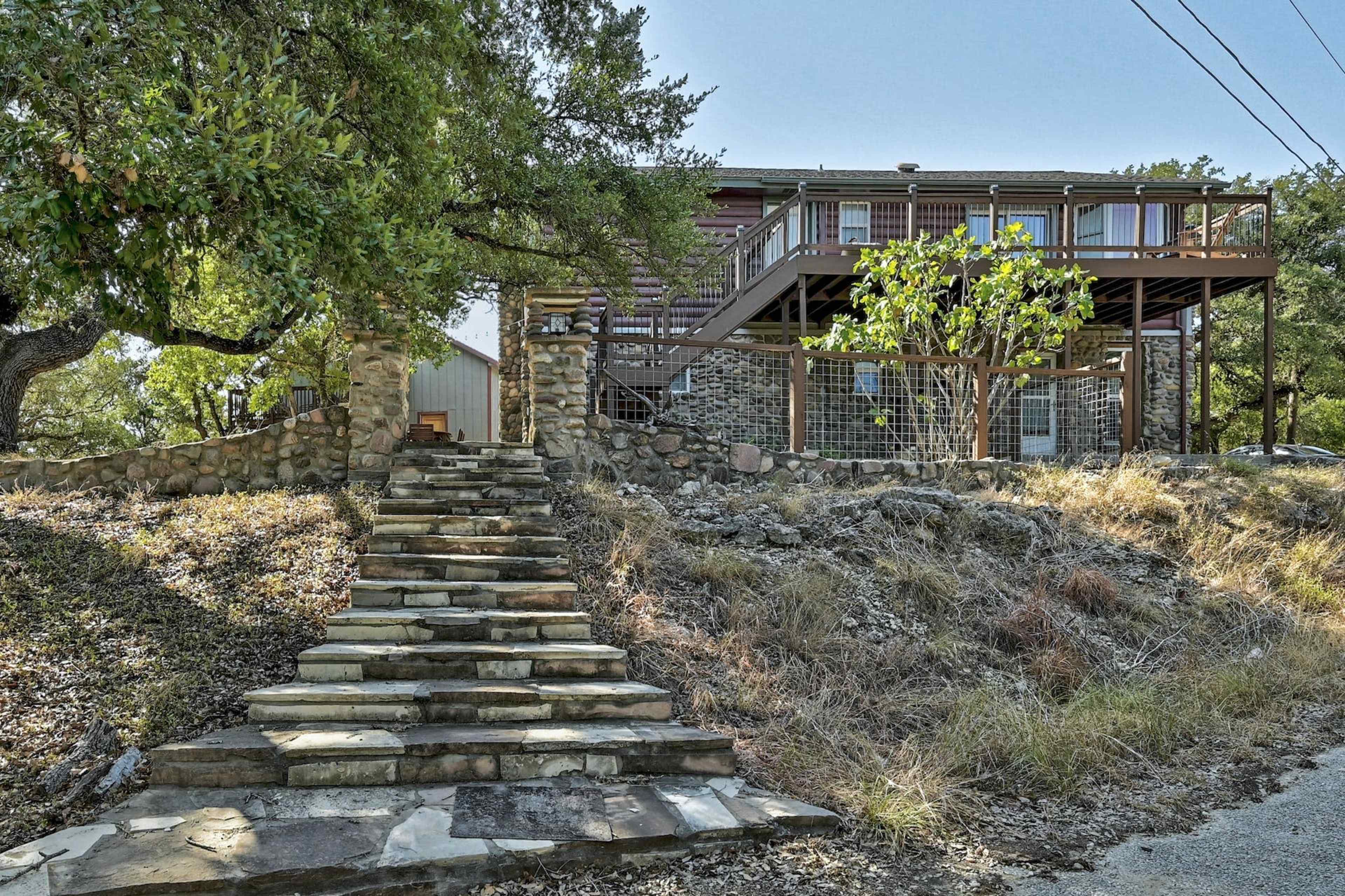A stone staircase leads up to a two-story house with a wooden deck, surrounded by trees and rocky terrain.